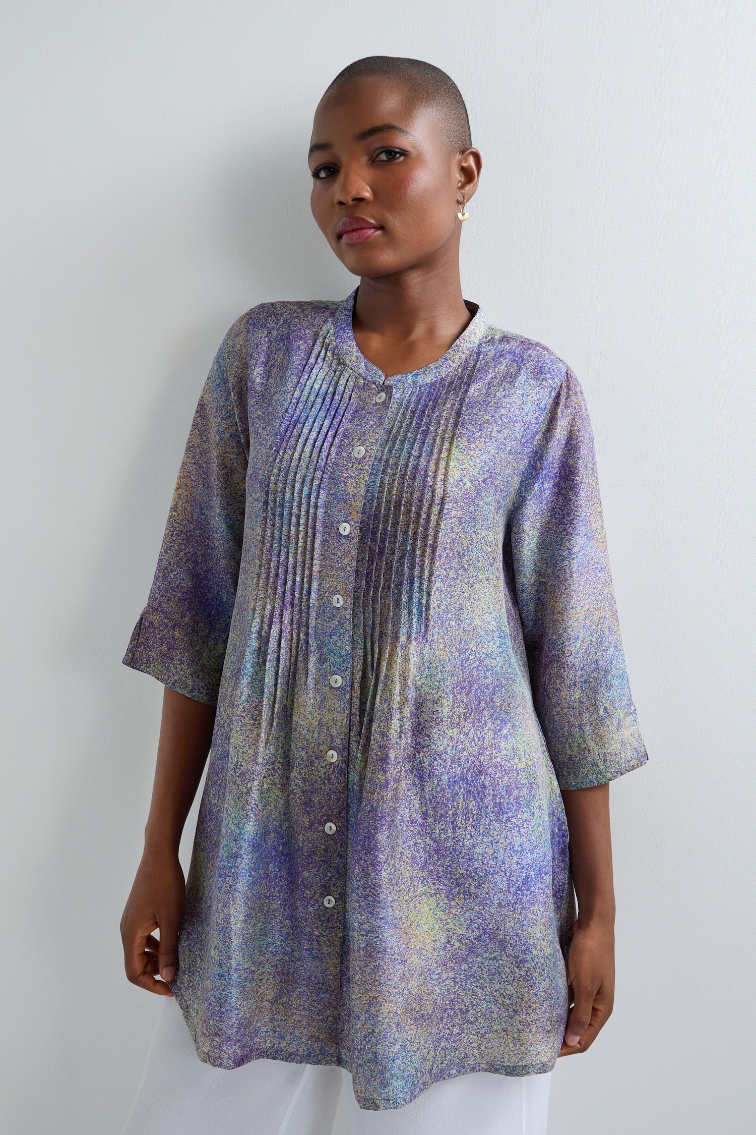 Against a plain light background, someone models the Abstract Speckled Silk Shirt—a pastel long-sleeve tunic with front pleats and buttons—showcasing its luxurious silk fabric and flattering fit-and-flare silhouette.