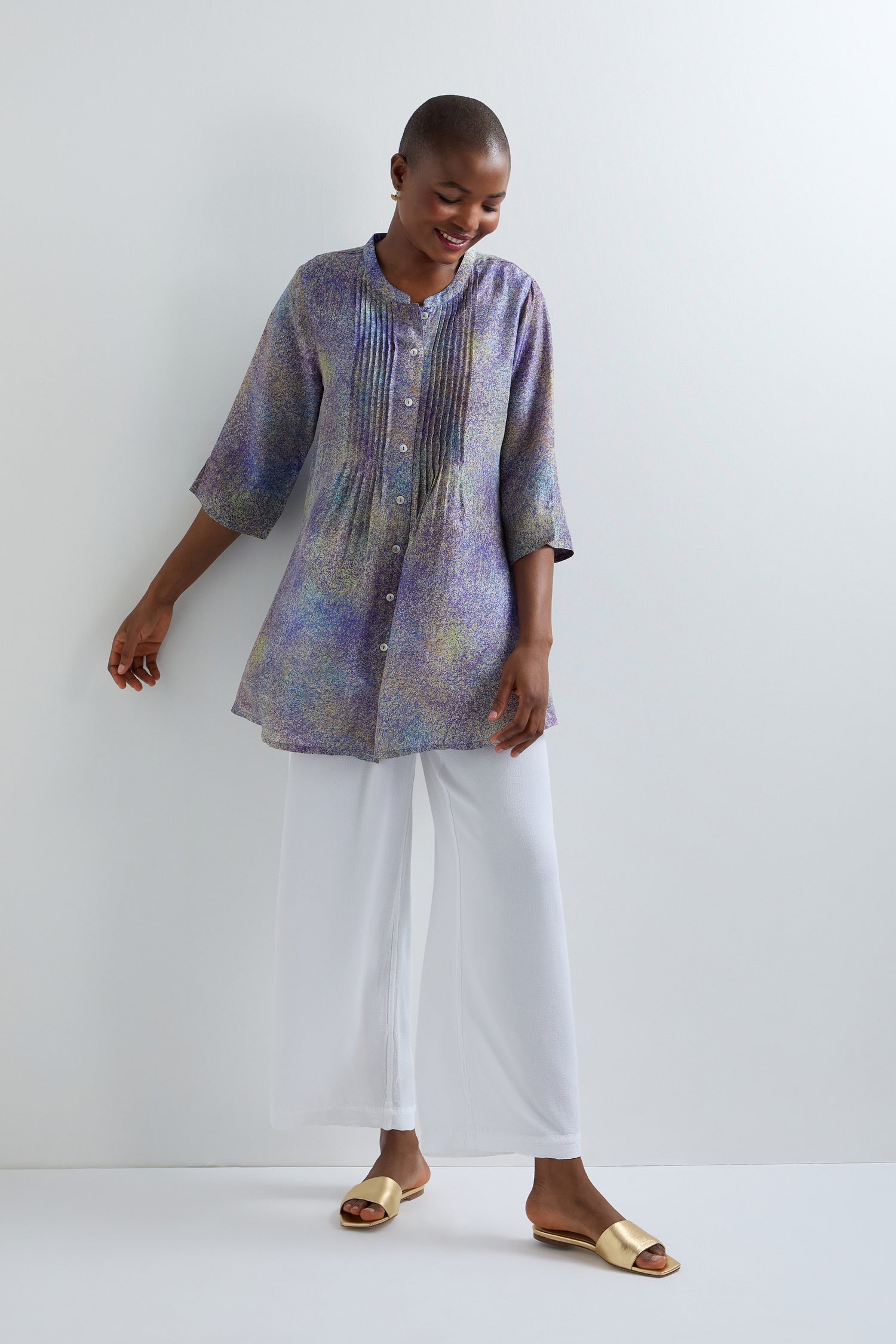 A person stands against a plain white wall wearing the Abstract Speckled Silk Shirt, a classic fit, long-sleeved button-up with pleats and stand collar, paired with white wide-leg pants and gold sandals.