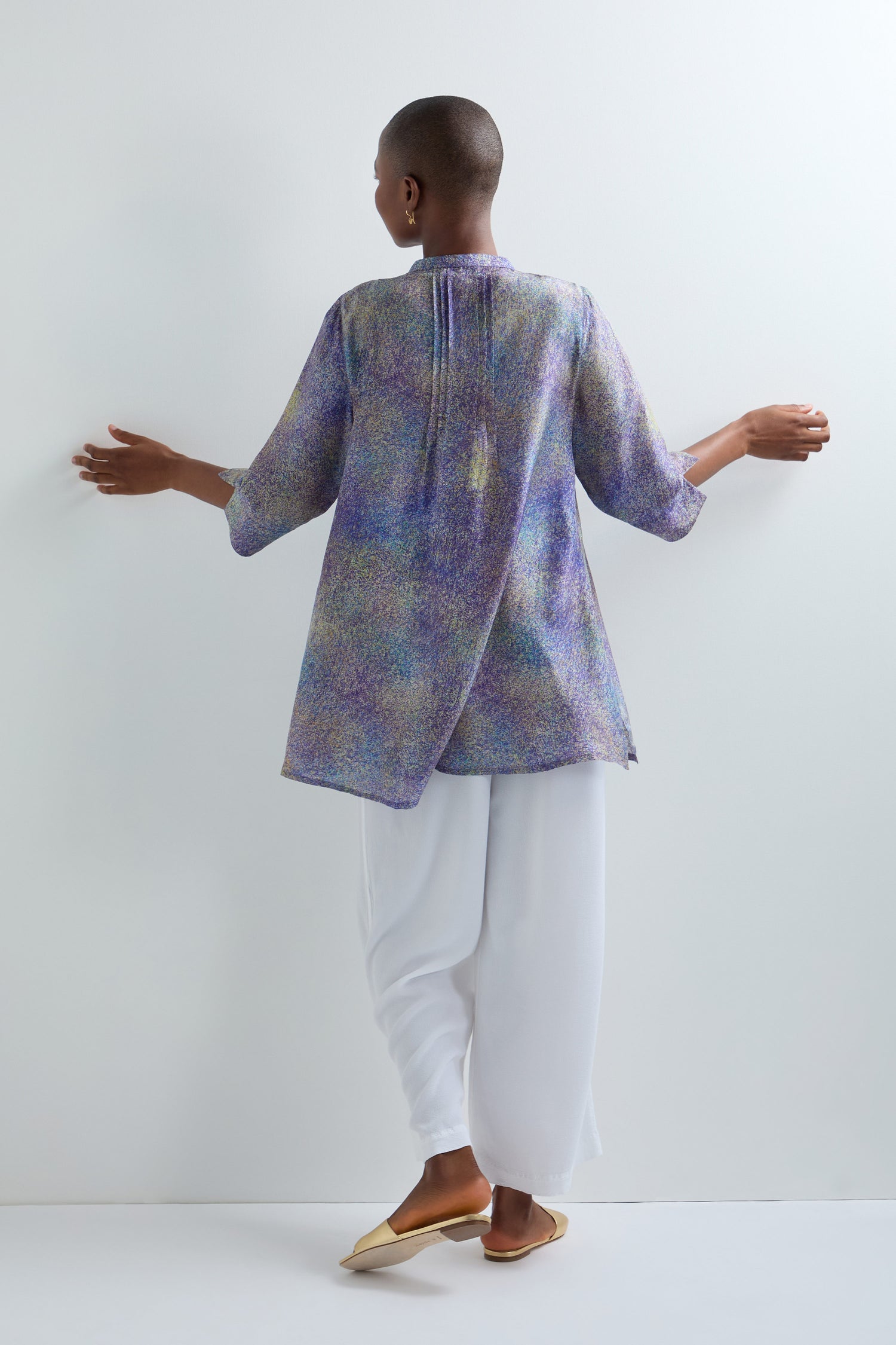 A person stands facing a wall, arms relaxed, wearing an Abstract Speckled Silk Shirt with a stand collar and long sleeves in a loose, multicolored style, paired with white wide-leg pants and tan flat shoes.