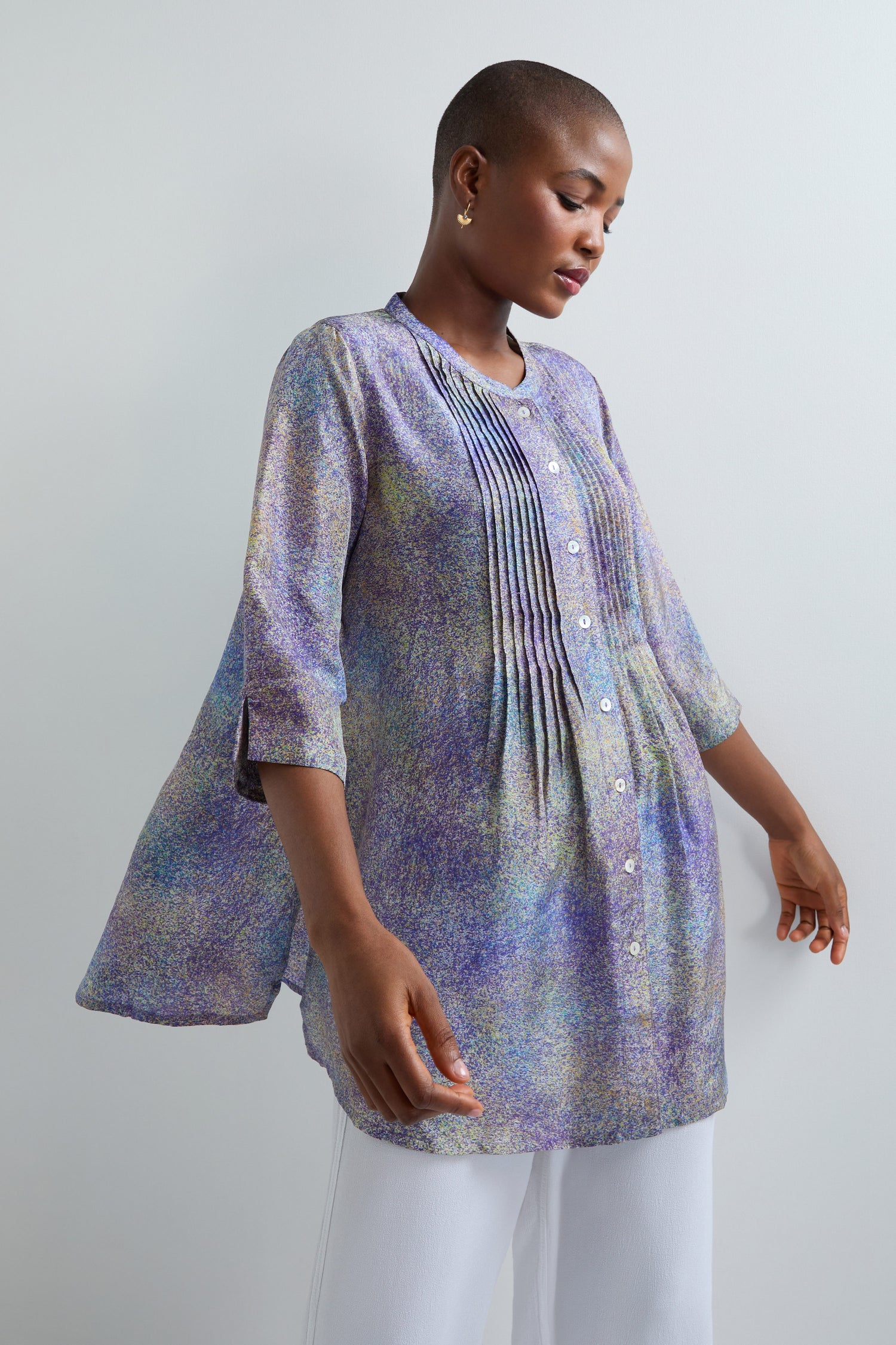 Someone in an Abstract Speckled Silk Shirt with long sleeves, pleats, a stand collar, multicolor pattern, and white buttons paired with white pants stands against a plain light background.