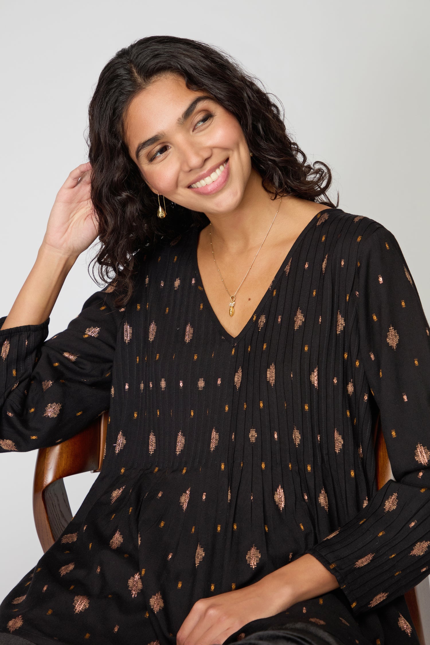 A woman with wavy dark hair sits on a chair, smiling and touching her hair while wearing the black Diamond Lurex Pleated Tunic and a gold necklace.