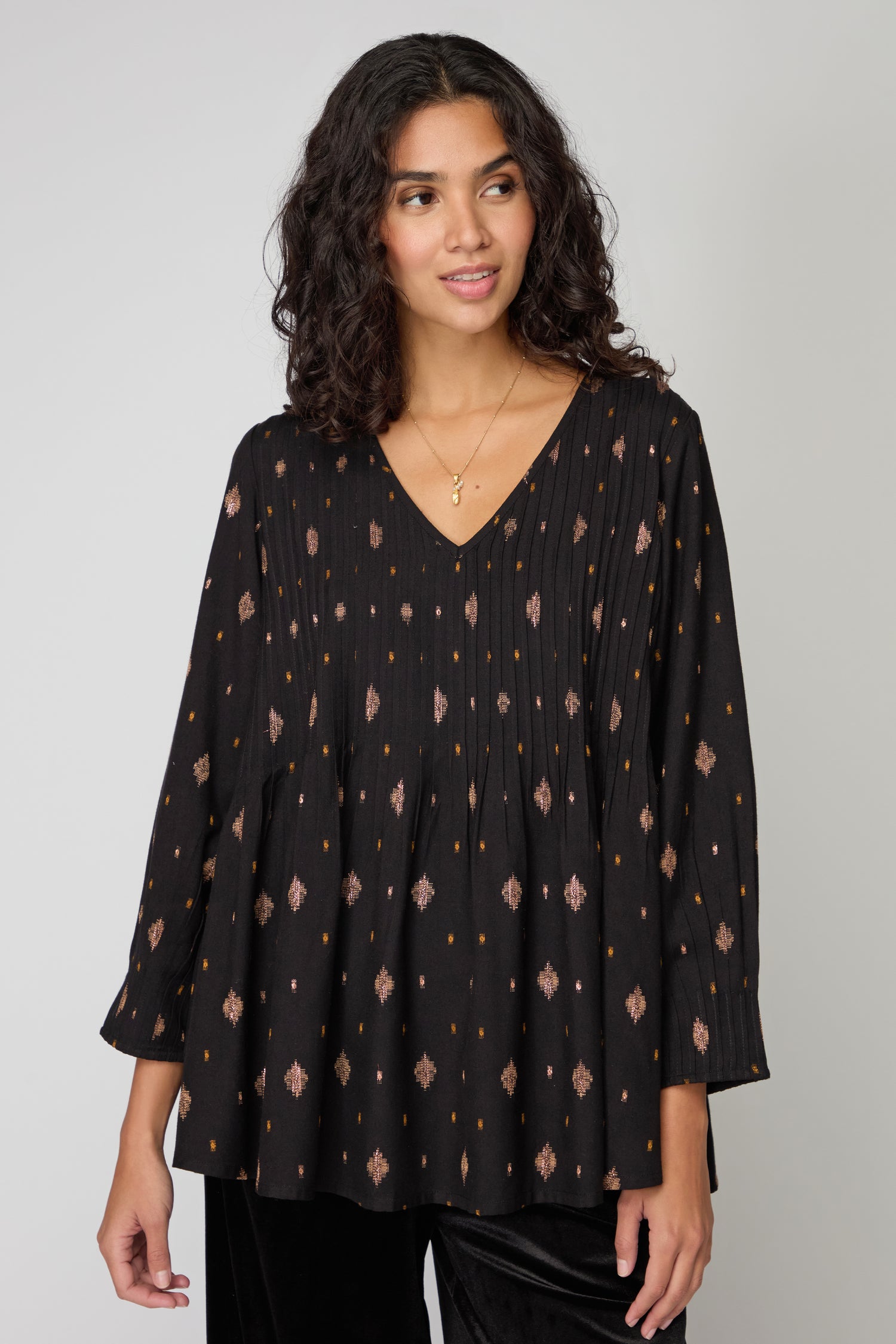 A woman wearing a loose-fitting black Diamond Lurex Pleated Tunic with a subtle geometric pattern and a V-neck stands against a plain light gray background.