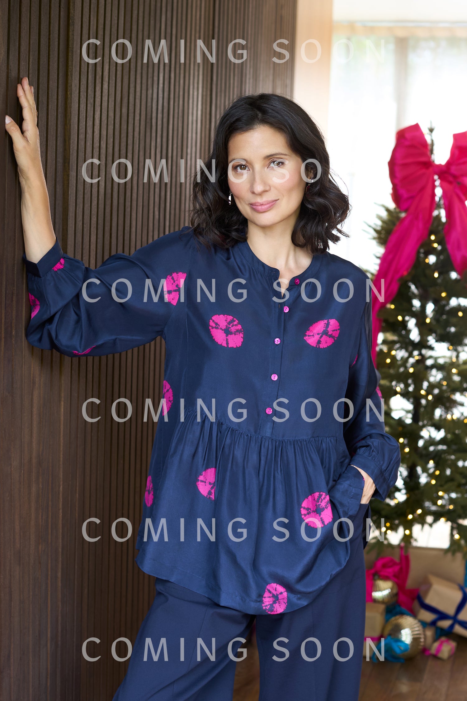 A woman wearing the Shibori Spot Silk Shirt in navy with pink prints stands indoors beside a Christmas tree adorned with pink bows.