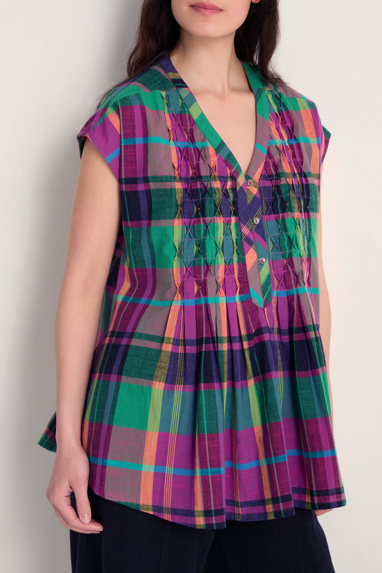 A woman wears the Madras Check Pleated Top, a pure cotton piece with short sleeves, V-neck, pleated front, and colorful green, purple, yellow, and pink check patterns.