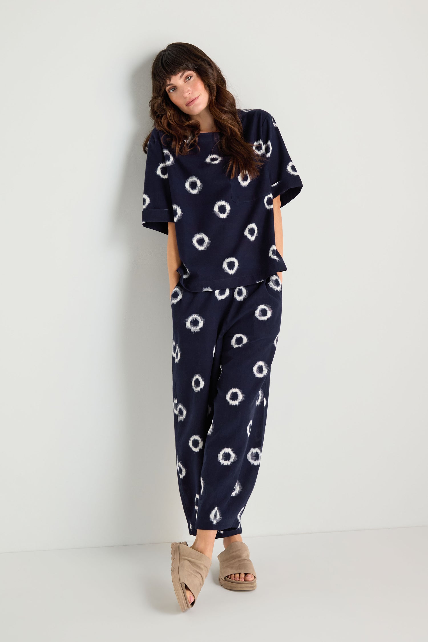A woman wearing the Ikat Circles Cotton Top, featuring a navy blue color, white circular patterns, and an oversized boxy silhouette, stands against a white wall with her hands in her pockets and beige sandals.