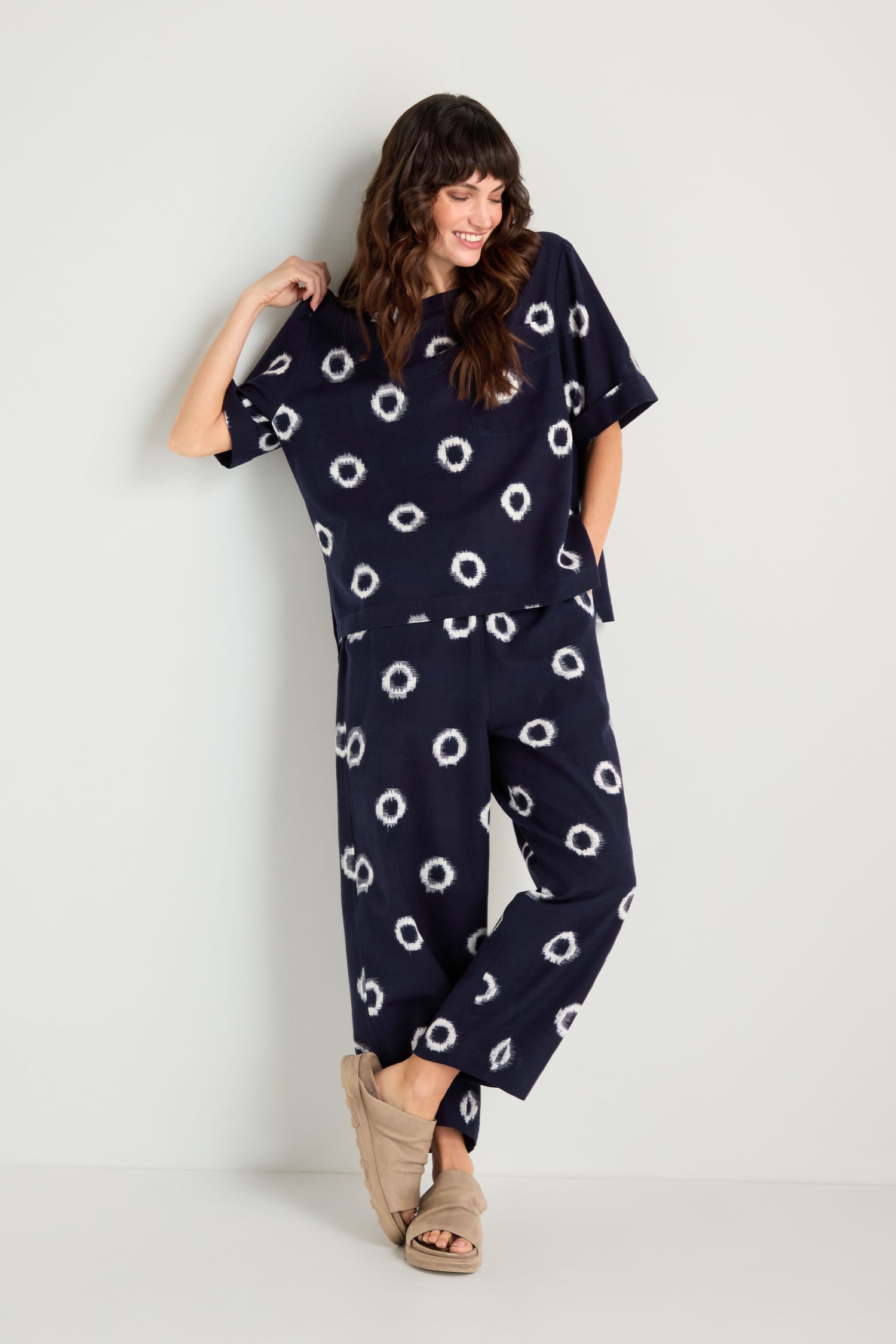 Woman standing against a white wall, wearing an Ikat Circles Cotton Top and matching pants in an oversized silhouette with white circular patterns, both crafted from double ikat cotton, tan slippers, looking to the side and smiling.