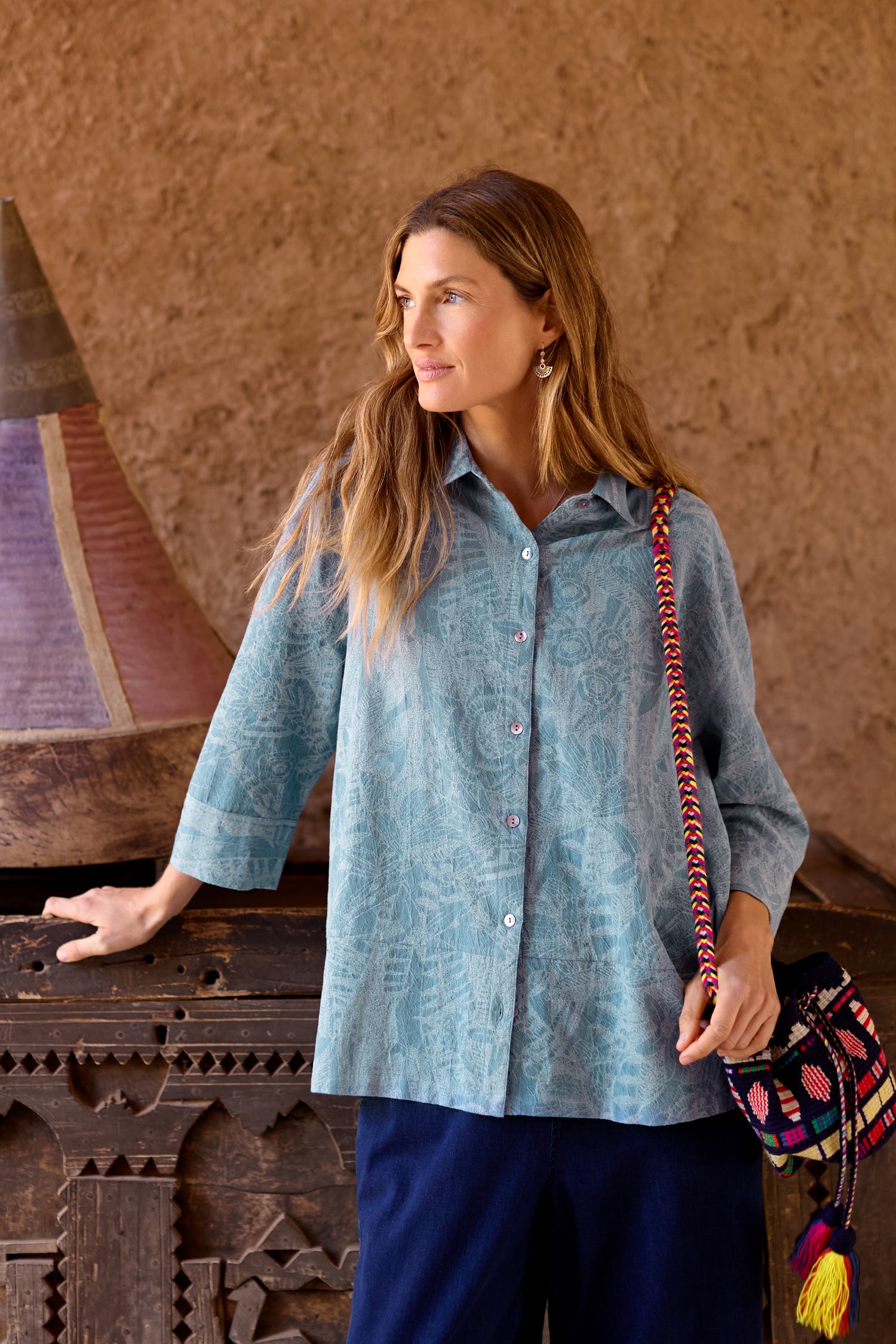 A woman stands indoors, wearing a Floral Jacquard Cotton Shirt and dark pants, with her hand on a carved wooden surface and a colorful GRT6856-FJC bag over her shoulder.