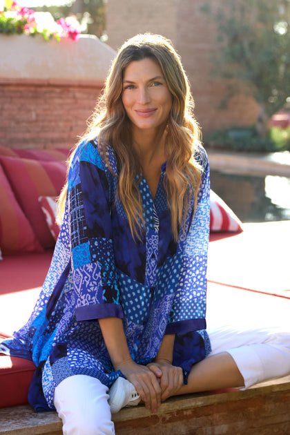 A woman with long, wavy hair sits cross-legged on a red outdoor sofa, smiling at the camera in the Sahara Indigo Patchwork Longline Shirt and white pants.