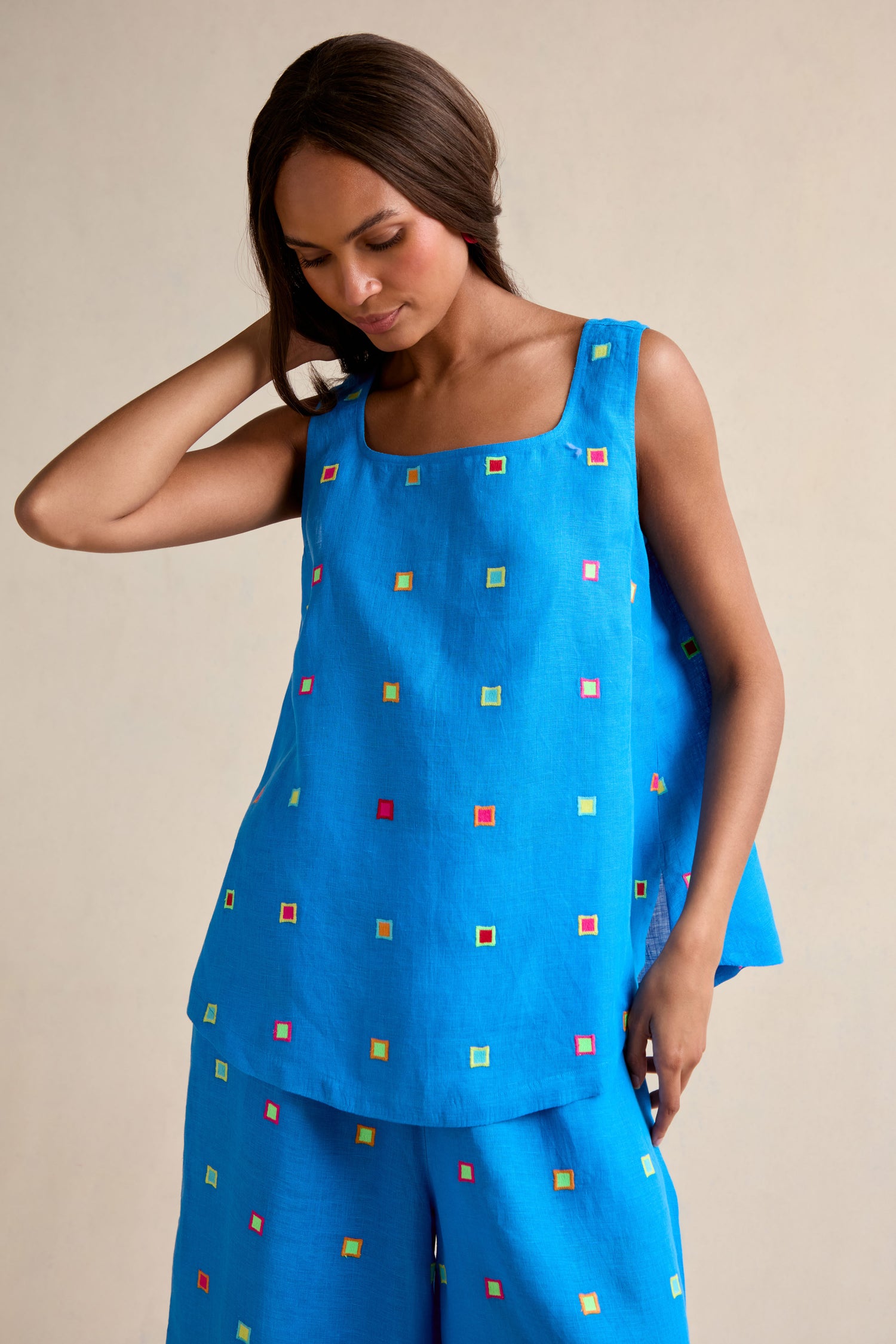 A woman models the Embroidered Squares Linen Vest, a relaxed fit blue sleeveless top with hand-finished multicolored square embroidery, paired with matching wide-leg pants, against a plain background.