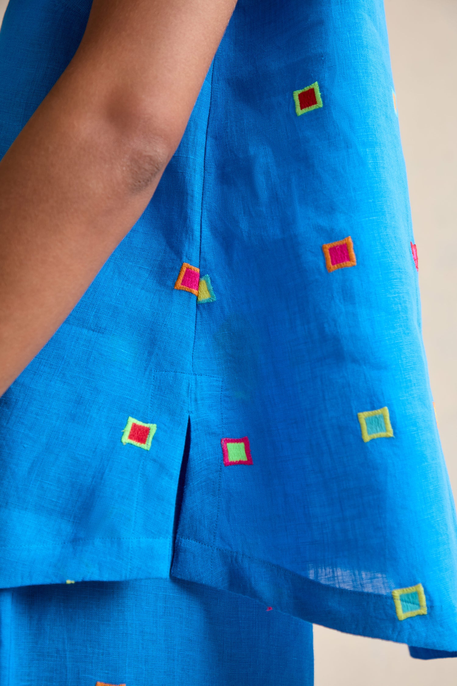 A person wears the Embroidered Squares Linen Vest, a relaxed fit, bright blue sleeveless top featuring multicolored hand-finished square embroidery and a side slit.