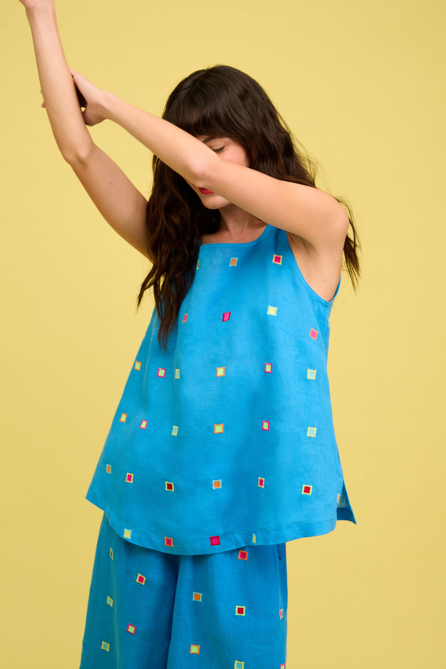 Woman wears the Embroidered Squares Linen Vest and matching pants with multicolored square patterns, standing against a yellow background with her arms raised.