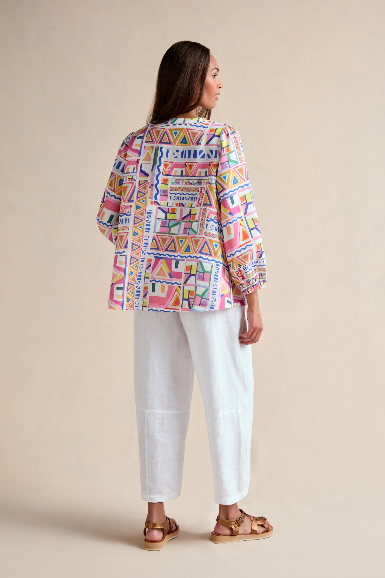 A woman stands facing away, wearing the Painted Geo Linen Top with balloon sleeves, paired with white ankle-length pants and brown sandals, set against a neutral background.