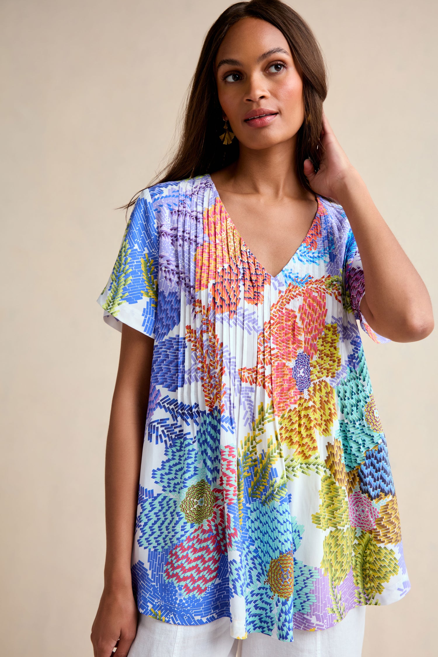A woman stands against a plain beige background, wearing the Pixelated Petals Print Top—a short-sleeved, V-neck top featuring a pixelated floral print and a relaxed A-line fit.