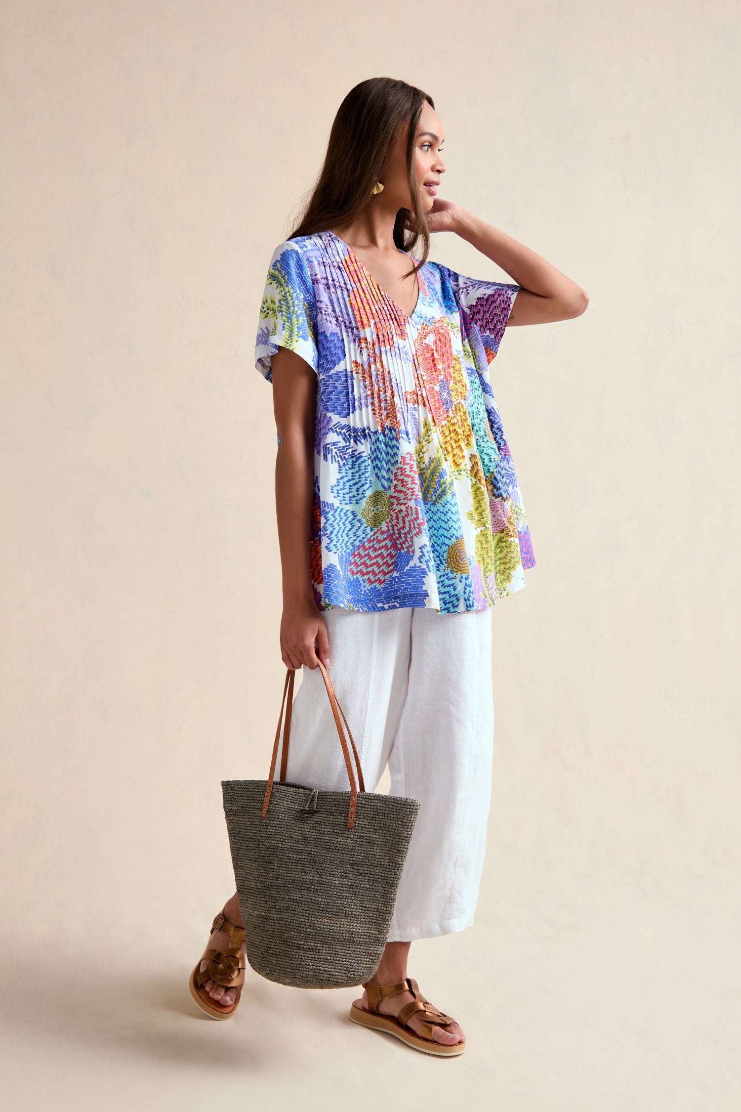 A woman stands holding a woven tote bag, wearing the Pixelated Petals Print Top with white wide-leg pants and brown sandals against a neutral background.