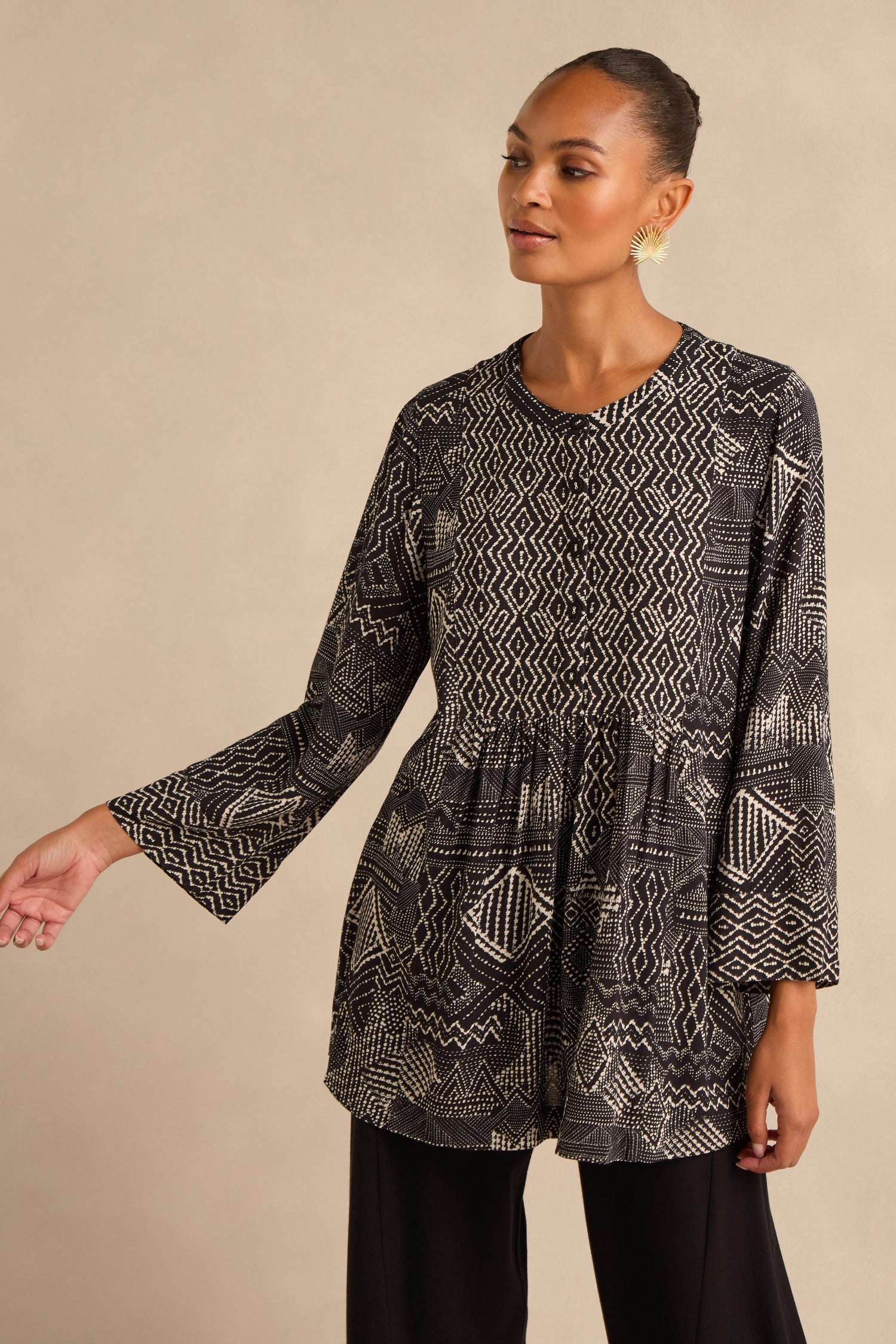 A woman wearing the Mono Patchwork Ikat Tunic, a long-sleeve black and white top featuring geometric patterns crafted from soft viscose crepe, paired with black pants, stands against a neutral background.
