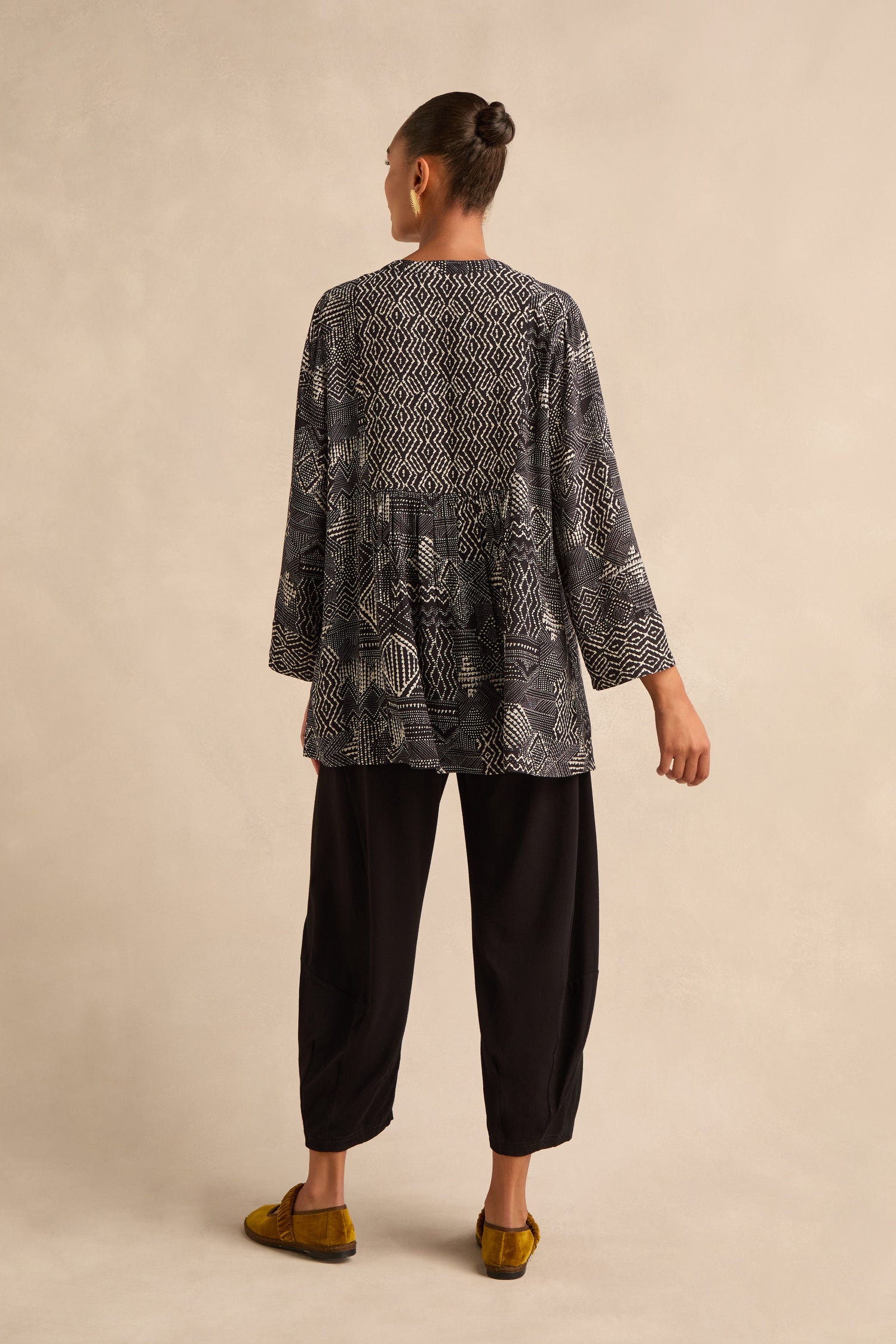 A woman stands facing away, wearing the Mono Patchwork Ikat Tunic crafted from viscose crepe in a black and white pattern, paired with loose black pants and mustard yellow flats, against a plain beige background.