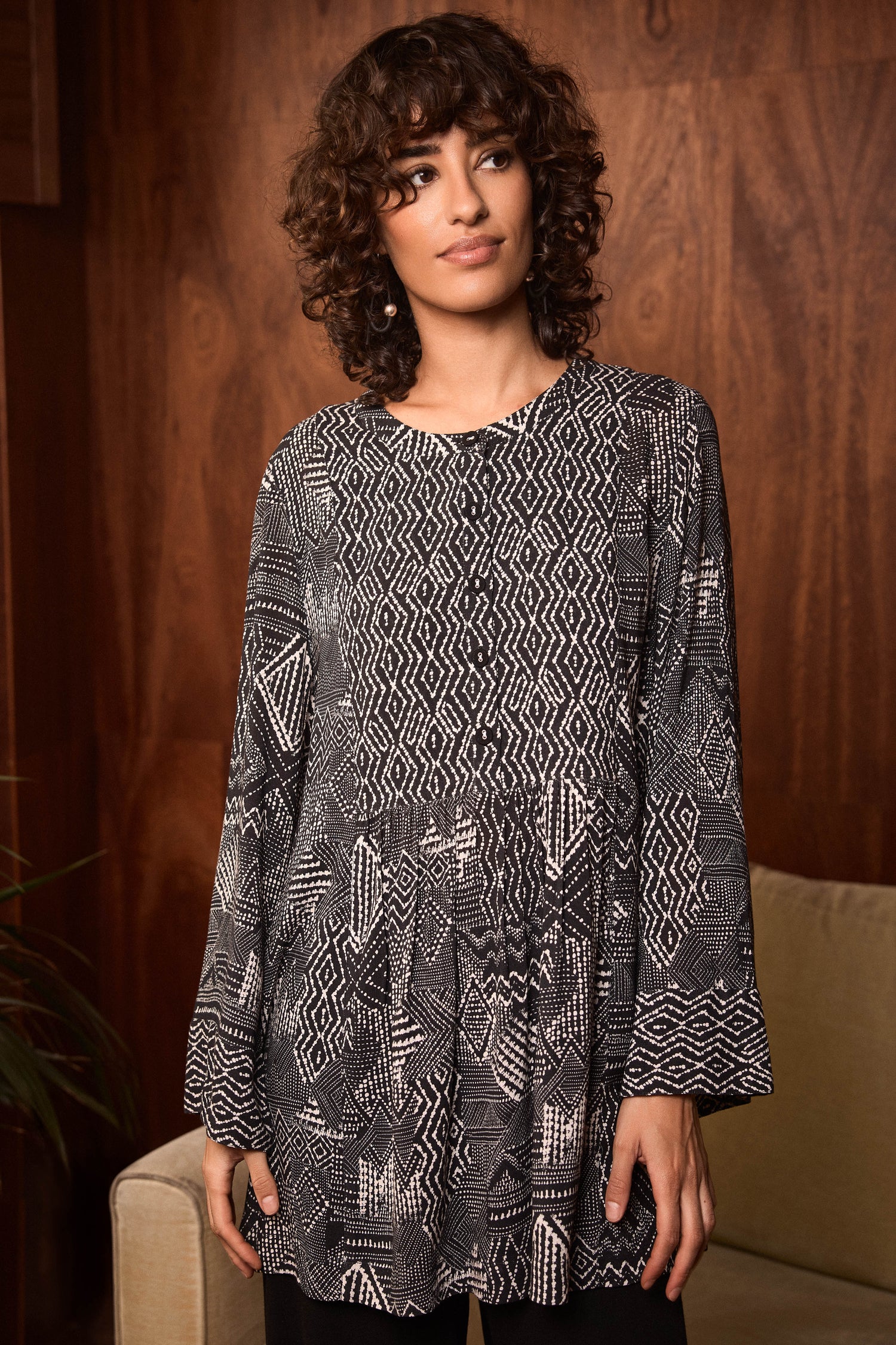 A woman with curly hair stands indoors in front of a wooden wall and beige sofa, wearing the Monochrome Batik Print Tunic, which features long sleeves.