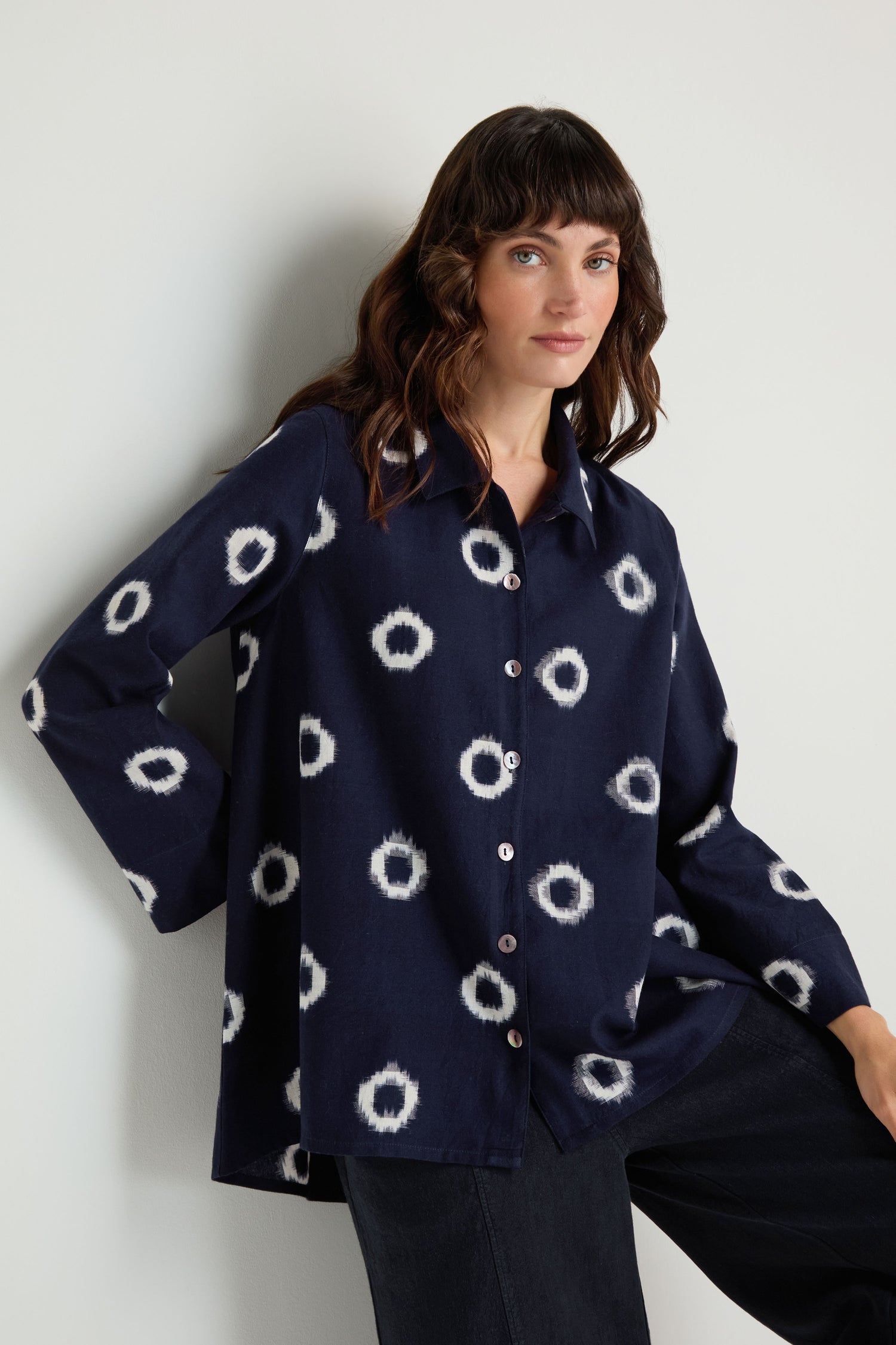A woman leans against a white wall with a relaxed expression, wearing the Ikat Circles Cotton Shirt in navy blue adorned with white circular patterns.