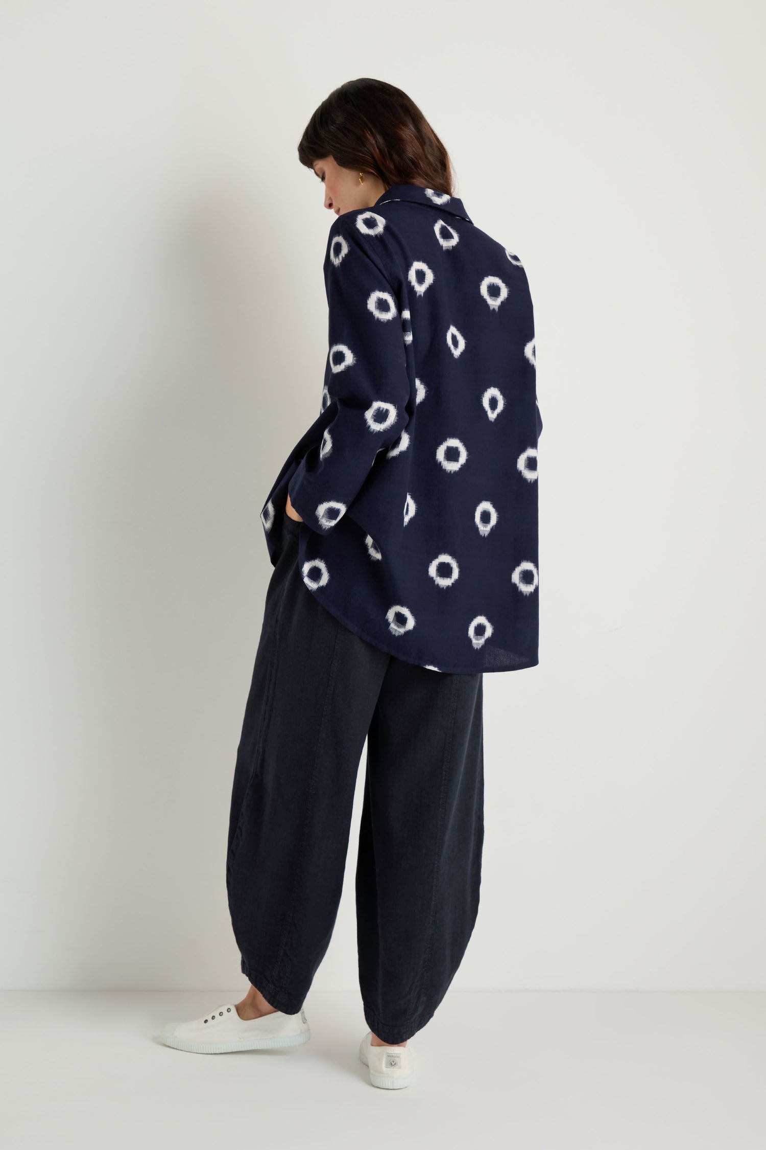 Person wearing the Ikat Circles Cotton Shirt—navy blue with white circular patterns—paired with dark loose pants and white sneakers, standing against a plain white background, facing away from the camera.
