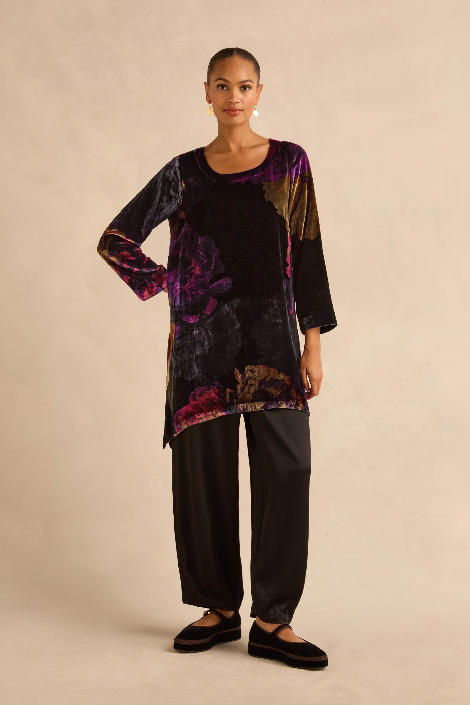 A person wearing a purple and gold Midnight Peonies Velvet Tunic paired with loose-fitting black trousers and black slip-on shoes.