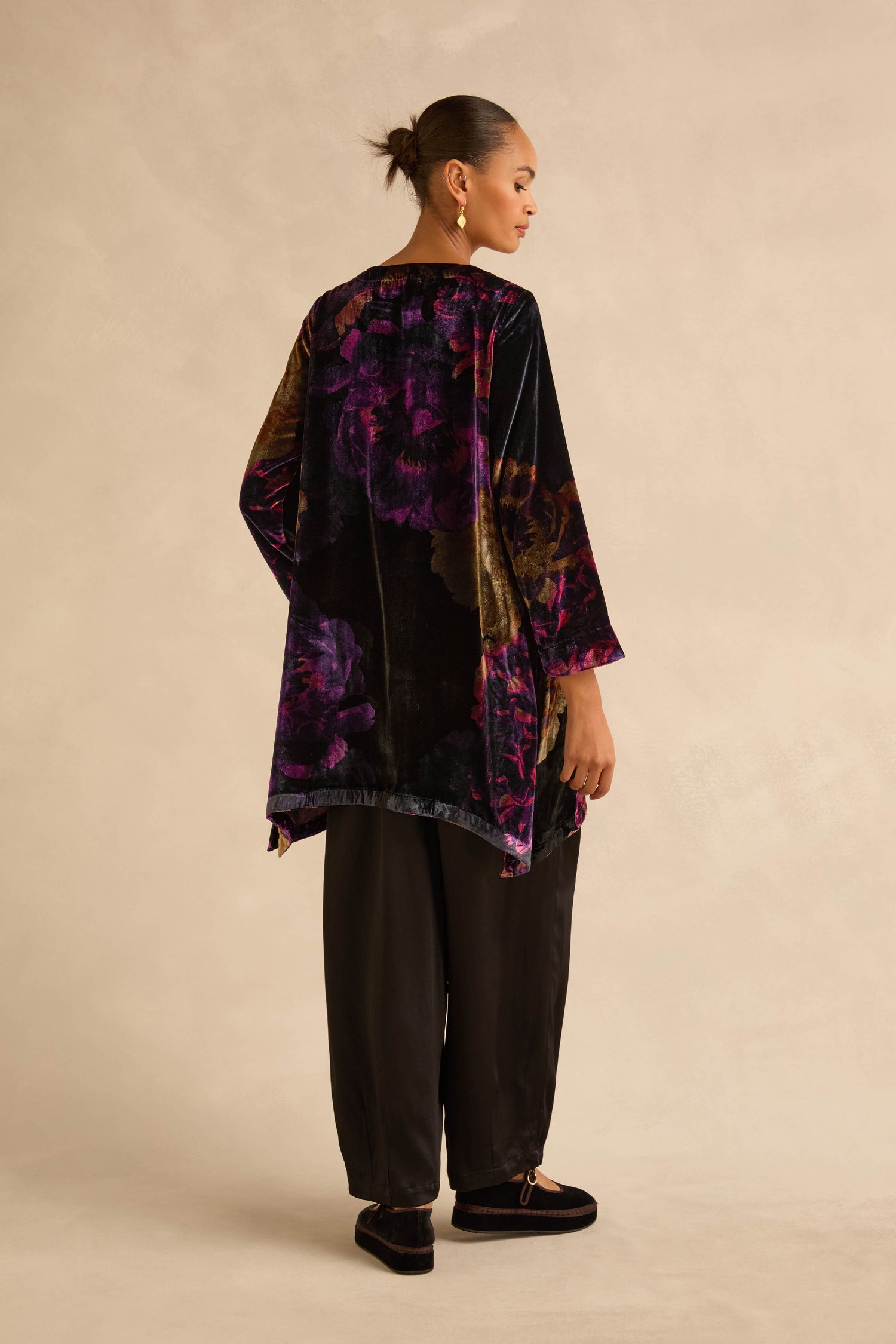 A person wears a Midnight Peonies Velvet Tunic in dark velvet with a purple and gold floral print, paired with loose-fitting black trousers and dark loafers.