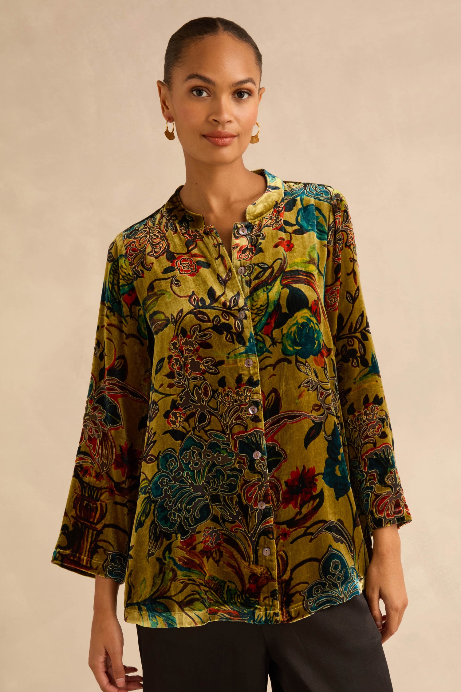 A woman wears a Silk Velvet Floral Devore Shirt, a long-sleeved, button-down velvet top featuring a multi-colored baroque floral pattern on a green base.