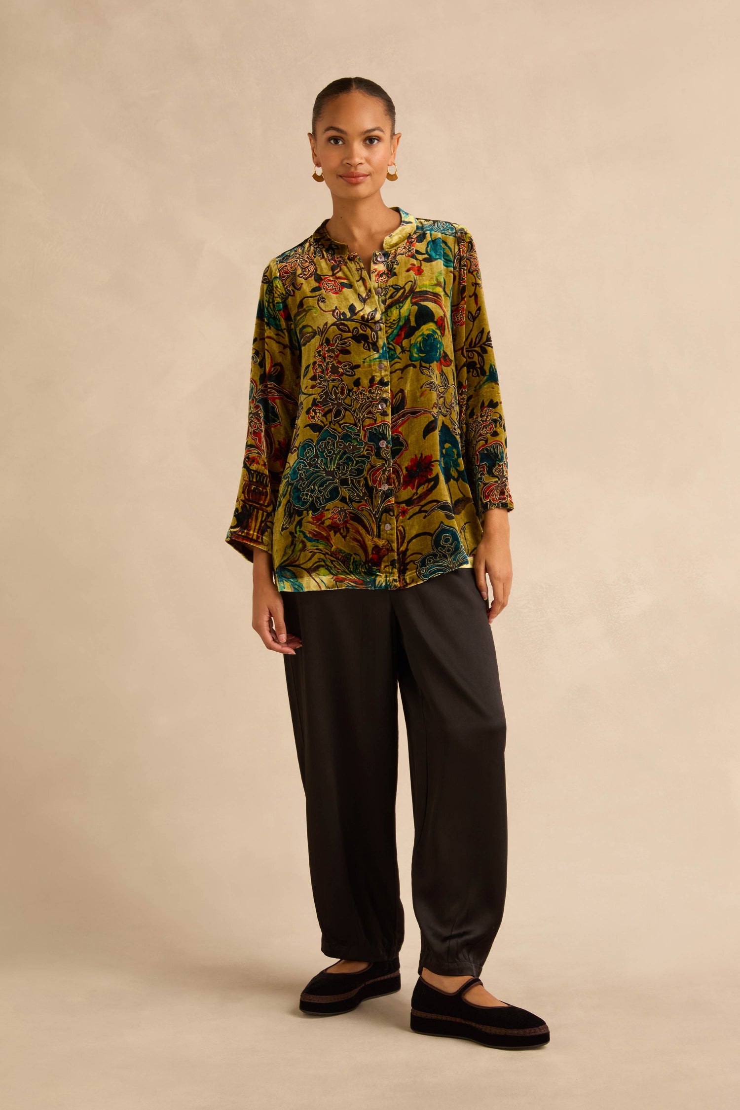 A person wearing a Silk Velvet Floral Devore shirt with a baroque pattern, loose-fitting black trousers, and black flats.