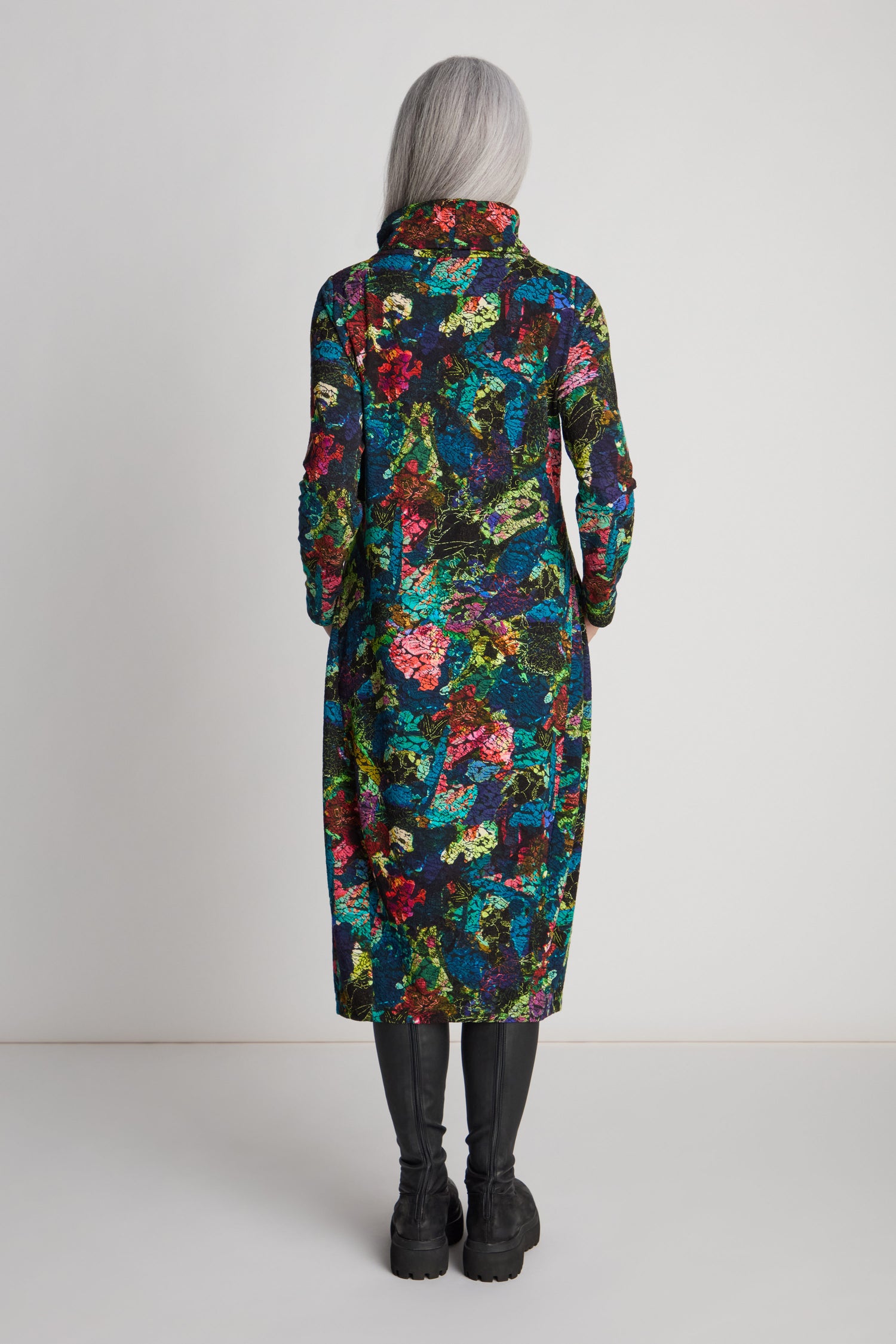 A person with long gray hair, viewed from the back, wearing a colorful Abstract Floral Jersey Bubble Dress and black boots, standing against a plain white background.