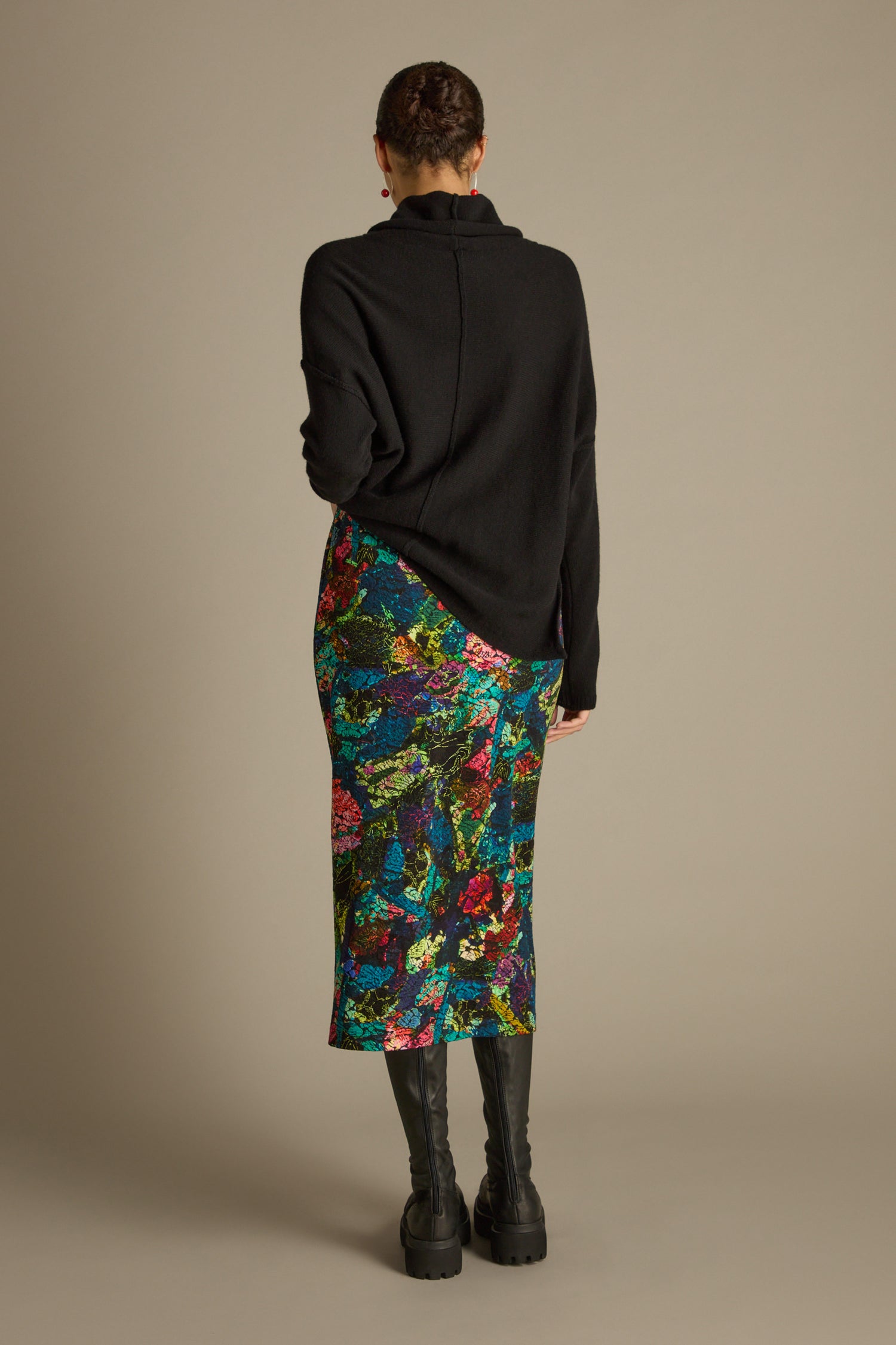 A person with short hair stands facing away, wearing a black sweater and the Abstract Floral Jersey Tube Skirt as a statement piece with black knee-high boots against a plain background.