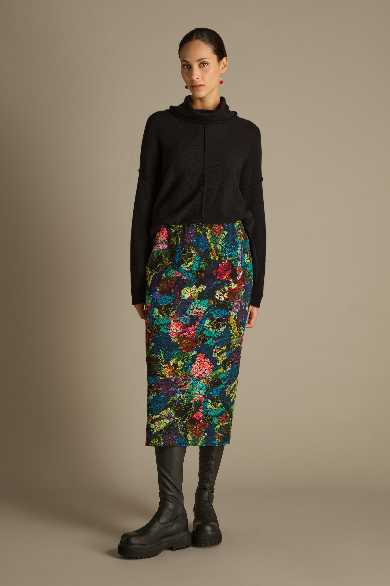 A person in a black sweater and the Abstract Floral Jersey Tube Skirt stands against a plain backdrop, wearing black boots.