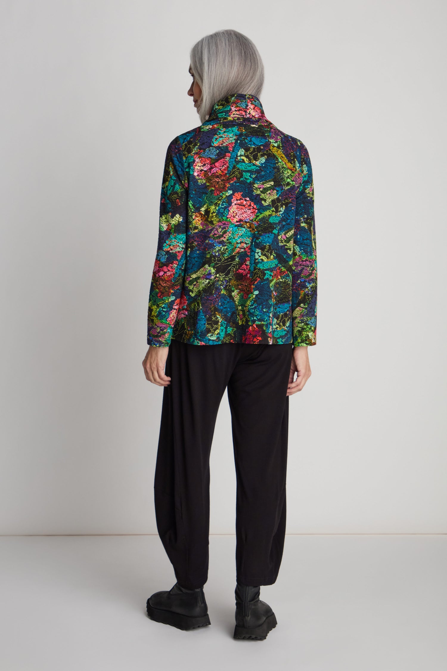 A person with long gray hair is standing, facing away, wearing an Abstract Floral Jersey Cowl Neck Top with a flared silhouette, black pants, and black boots.