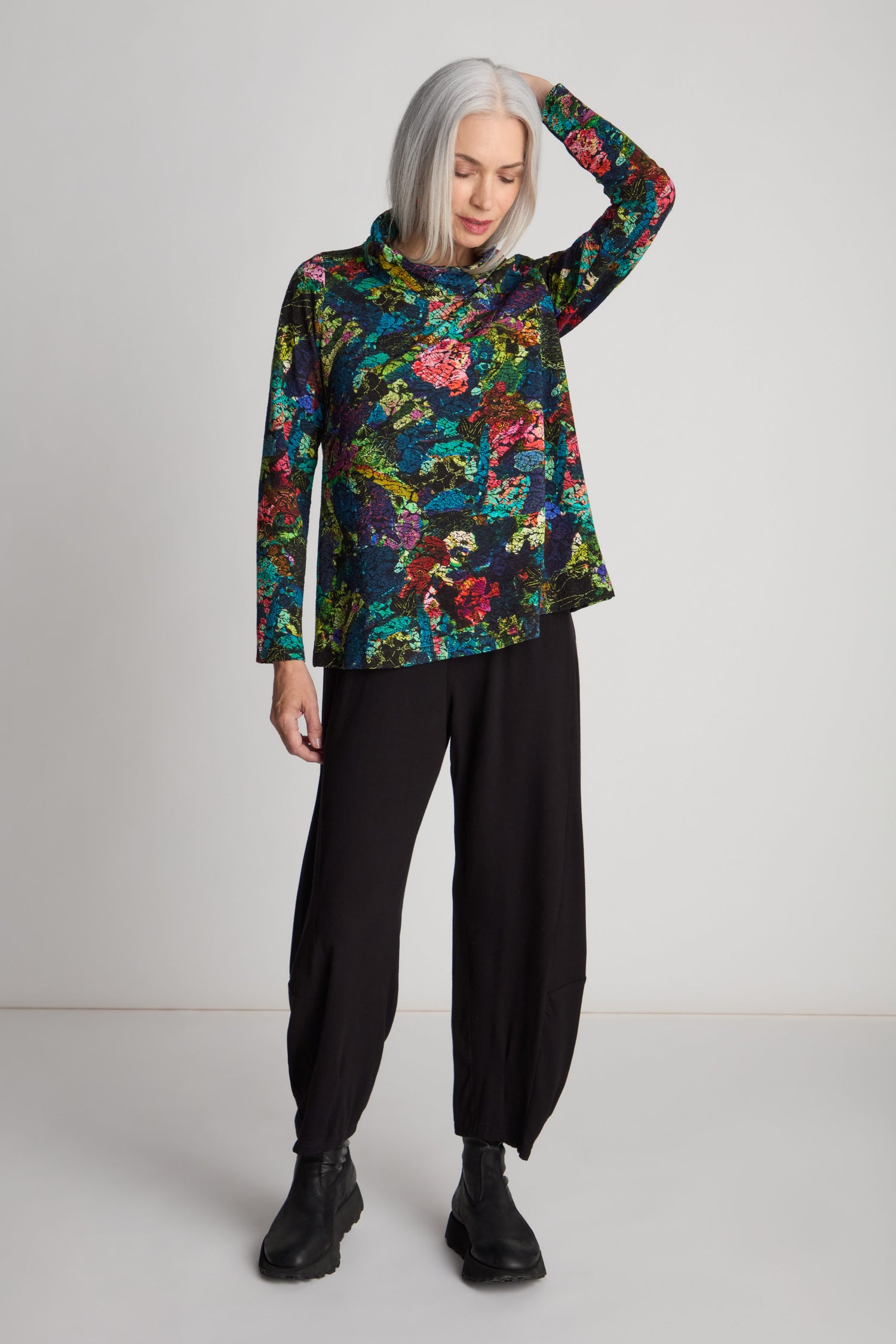 A woman with grey hair wears an Abstract Floral Jersey Cowl Neck Top with a flared silhouette, black loose pants, and black boots, standing against a plain background with one hand on her head.