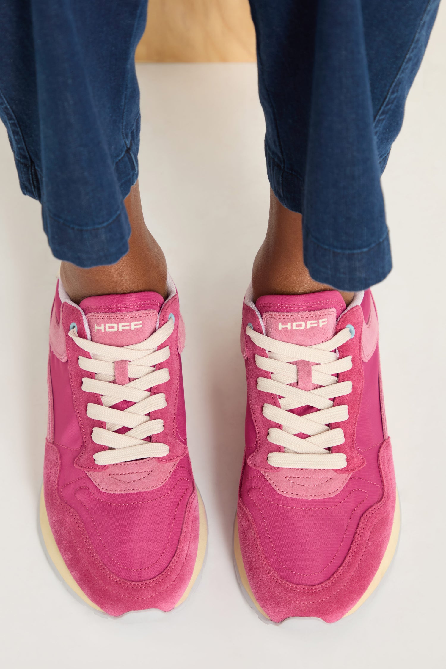 Someone wearing dark blue pants and City Retro Trainers in bright fuchsia with white laces stands on a light-colored surface.