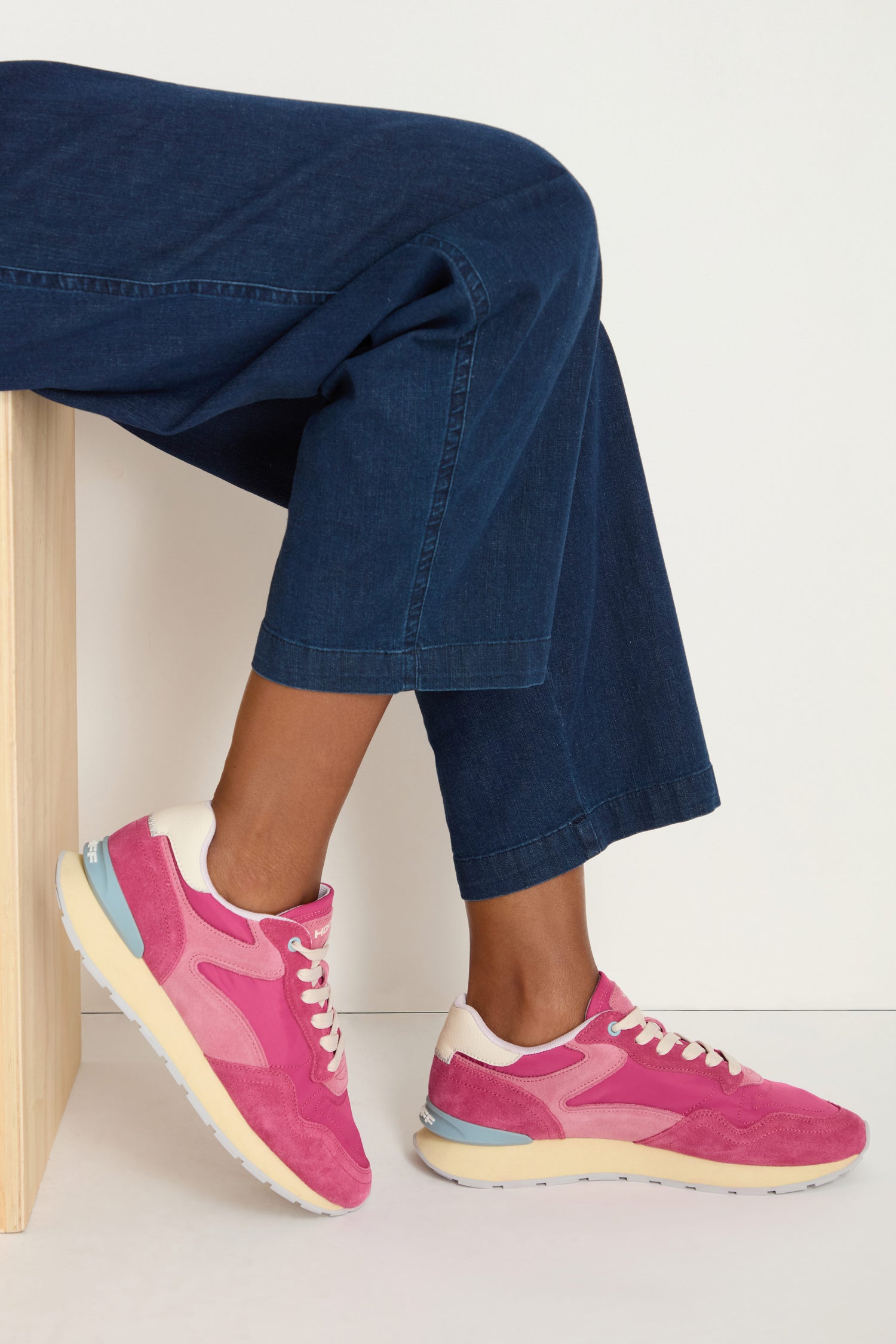 Someone sits on a light wooden bench against a plain background, wearing cropped blue jeans and City Retro Trainers with a memory foam insole.