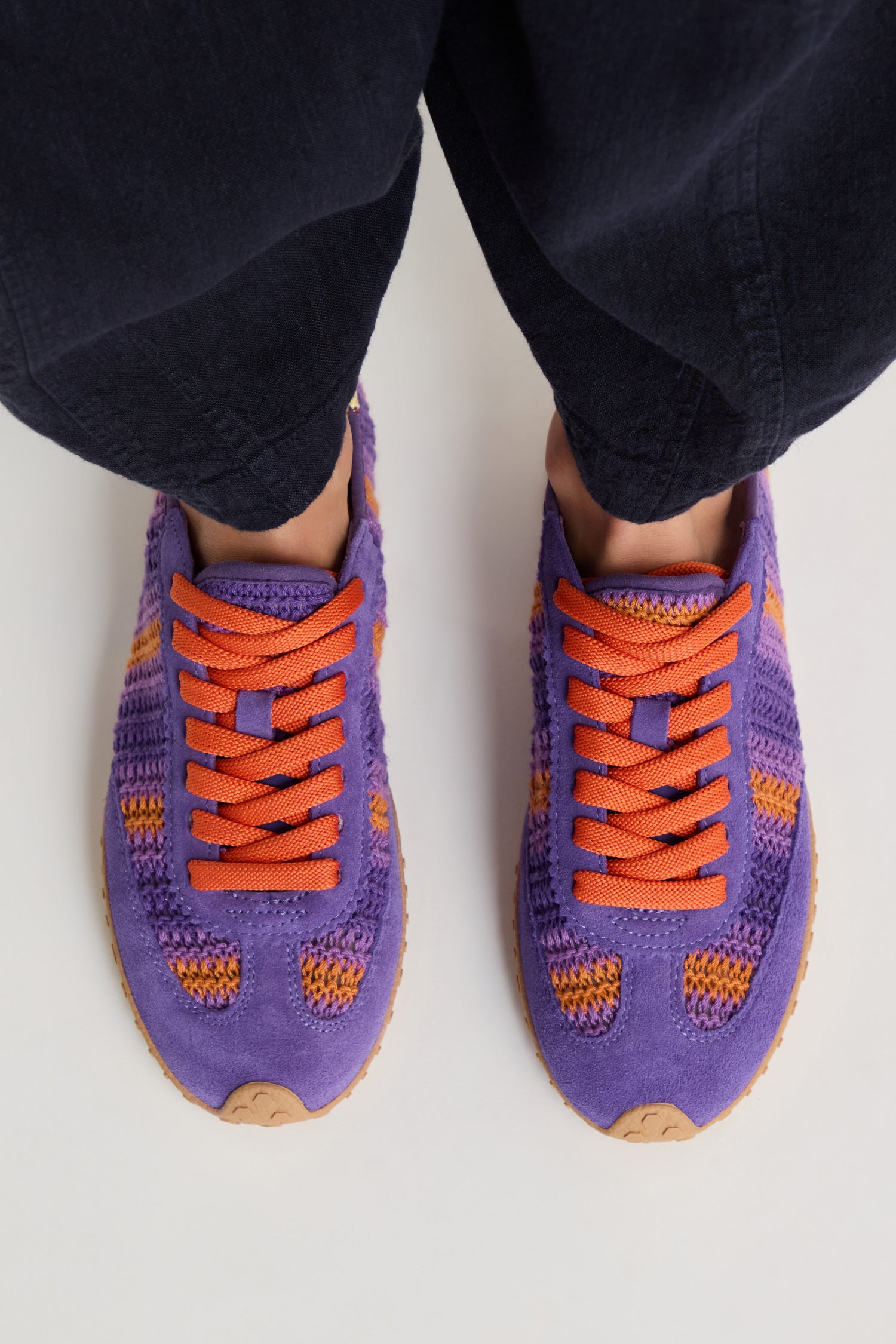 A person is wearing Bridge Woven Purple Crochet Trainers with orange laces, standing on a light surface. The black pants and the cushioned sole of the trainers are clearly visible from above.