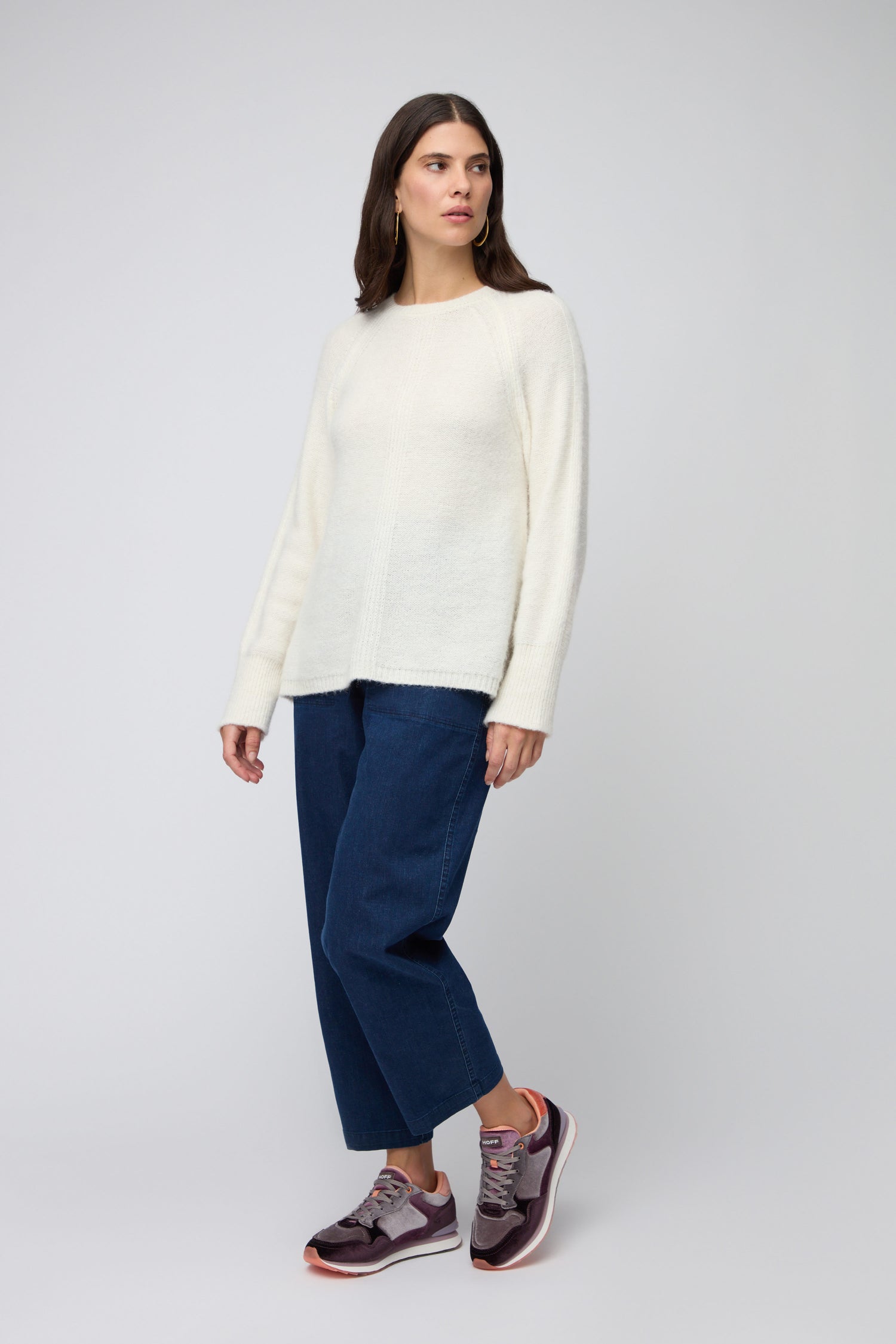 A woman faces left against a plain light background, dressed in a white sweater, blue wide-leg jeans, and Purple City Velvet Trainers.