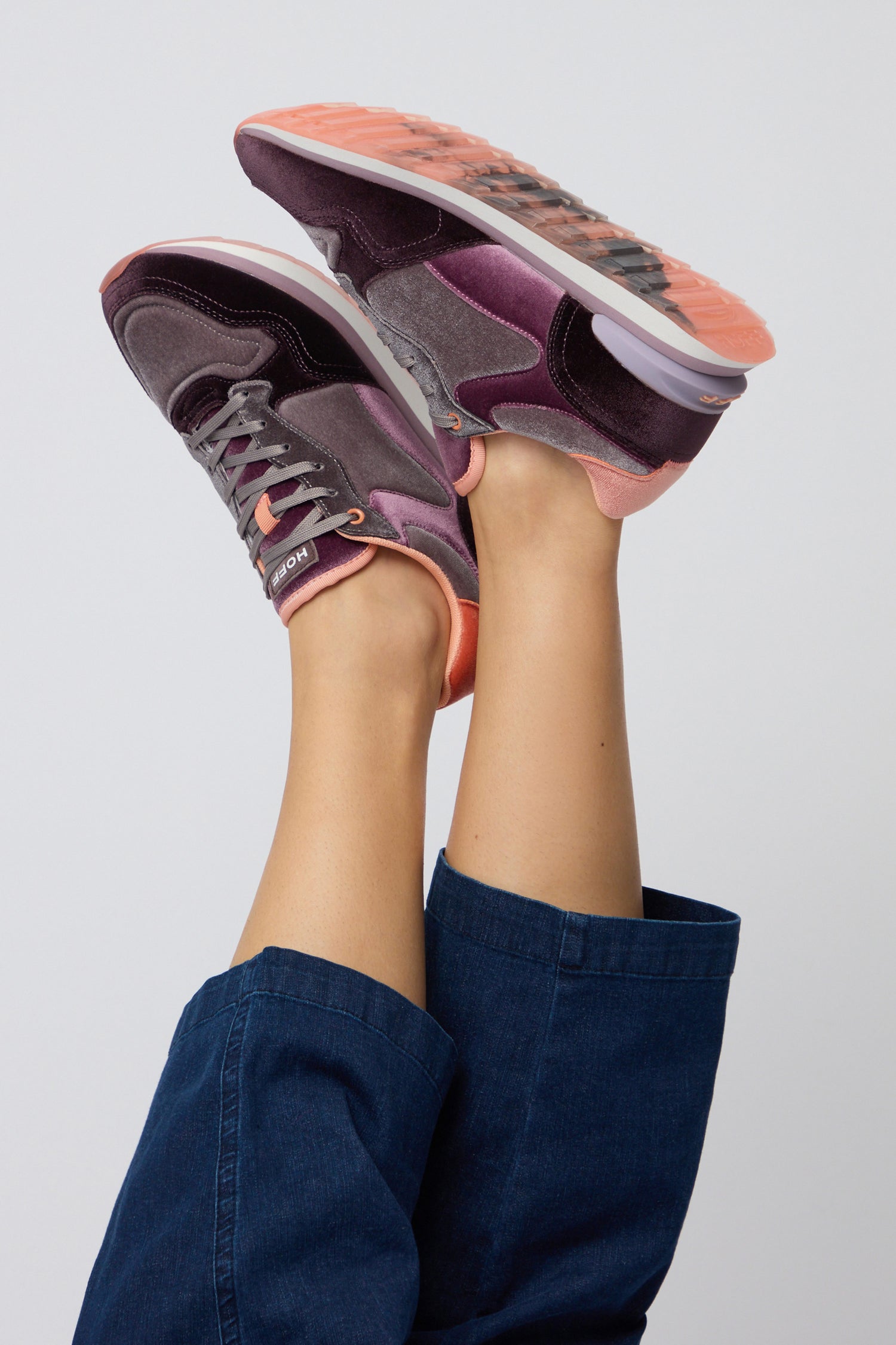 A person in cropped blue pants raises their legs, showing off the Purple City Velvet Trainers—women’s casual shoes in purple and pink with orange soles—against a plain background.