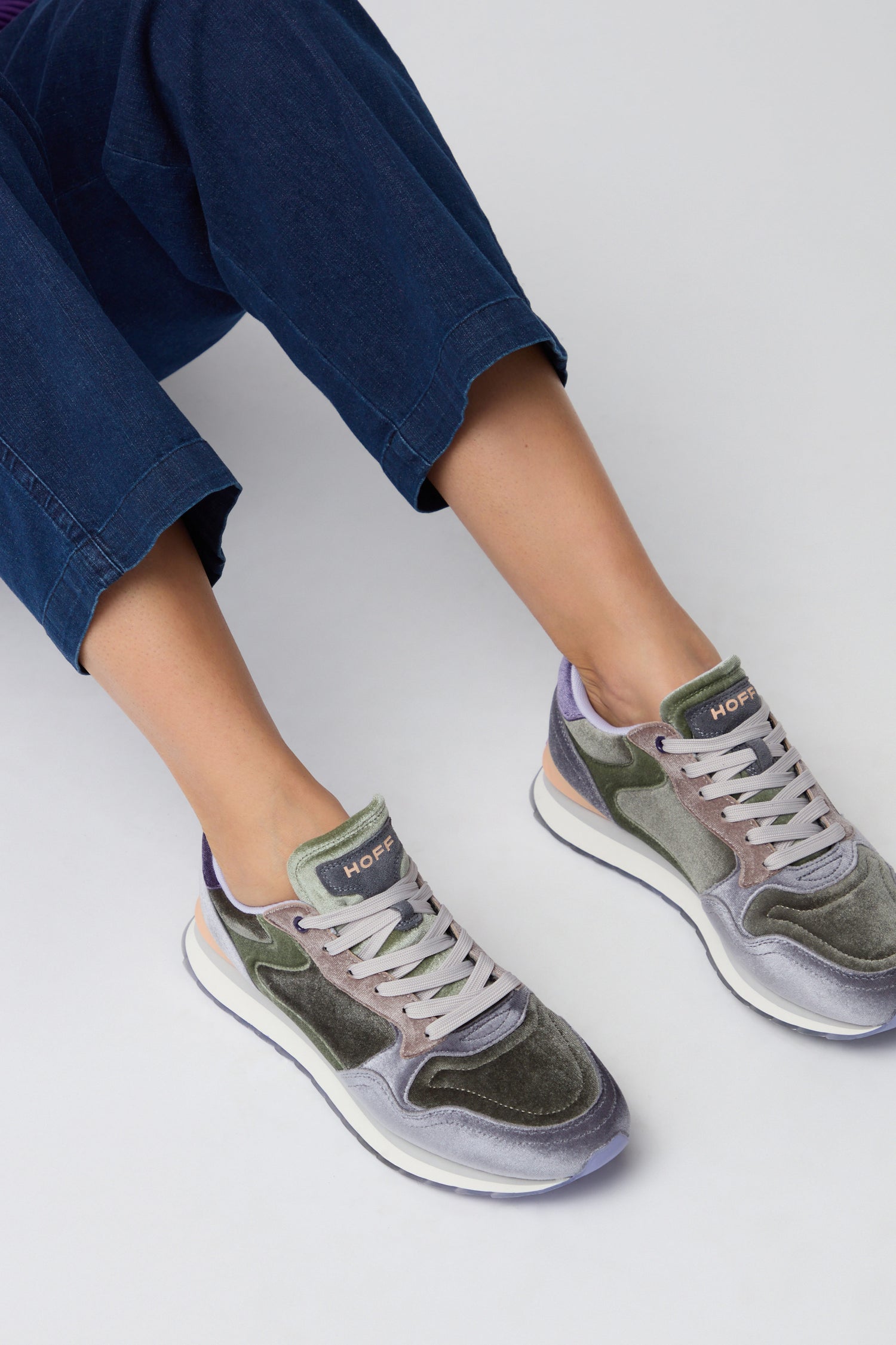 Person wearing blue pants and Green City Velvet Trainers, sitting against a plain light background.