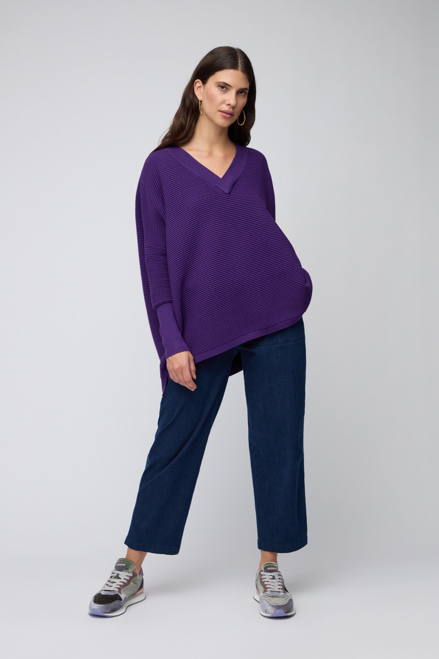 Woman standing against a light gray background, wearing a loose purple V-neck sweater, wide-leg blue jeans, and stylish Green City Velvet Trainers.
