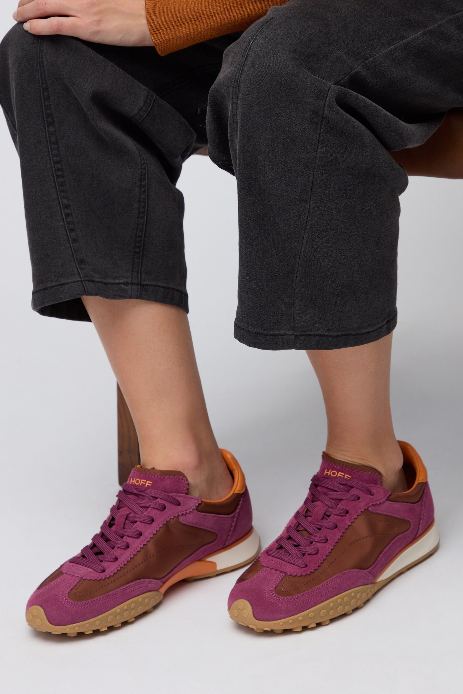 A person seated on a chair sports HOFF’s Burgundy Bridge Trainers with tan soles, paired with cropped dark gray jeans—an ideal choice for comfortable everyday women’s sneakers in purple and brown leather.