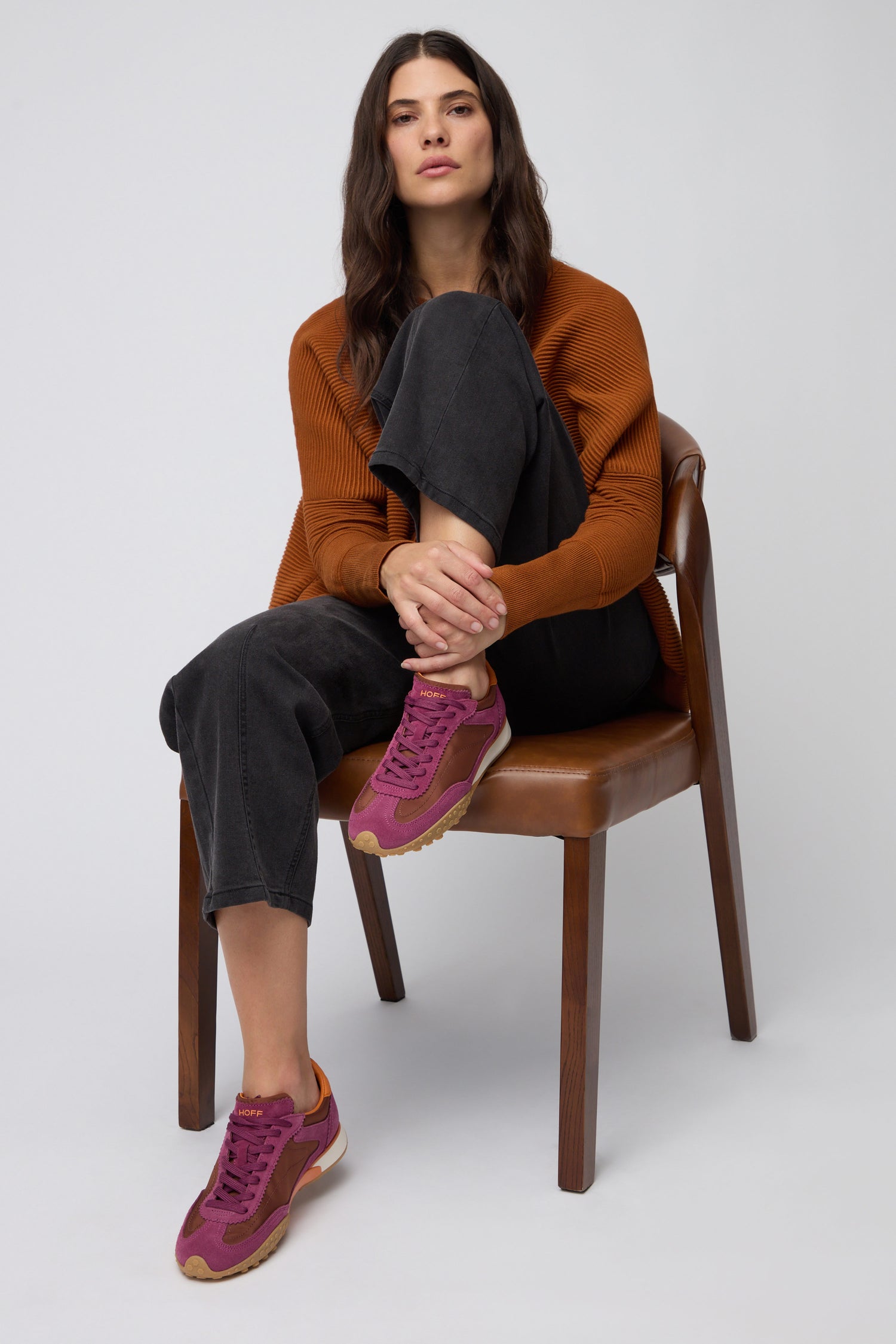 A woman with long brown hair sits on a wooden chair, wearing a rust sweater, black pants, and Burgundy Bridge Trainers, with one leg crossed over the other.
