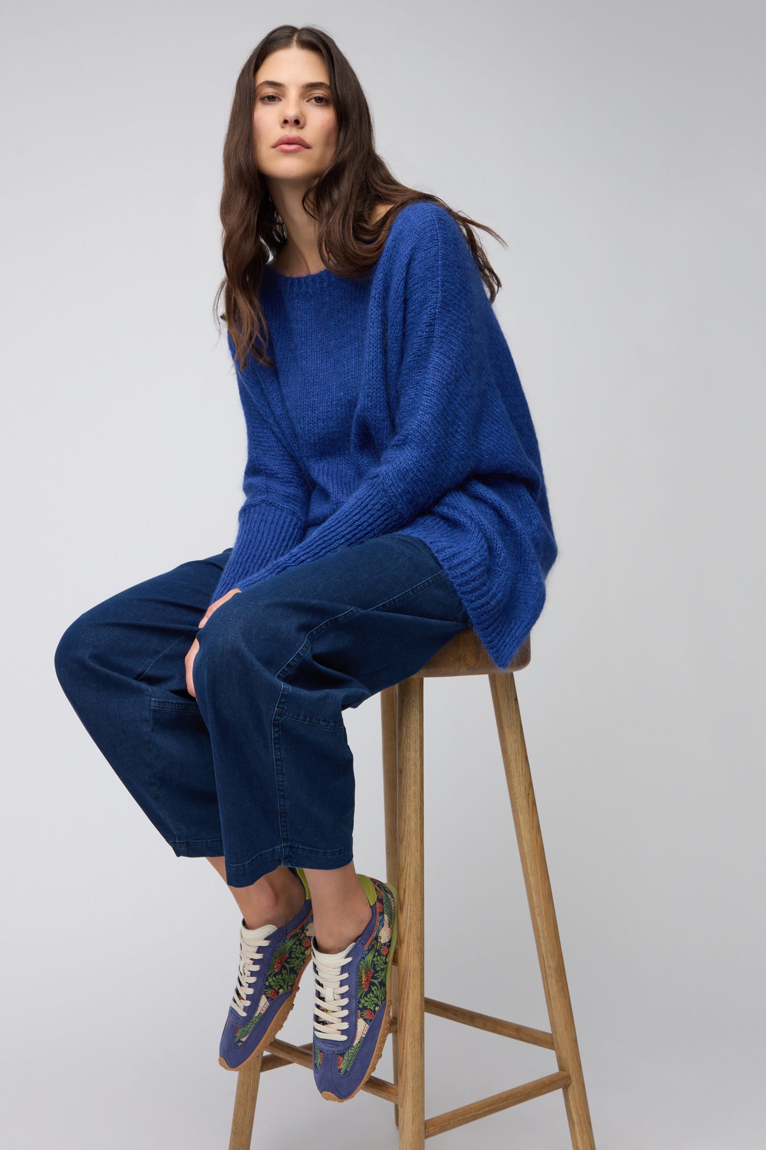 A woman with long brown hair, dressed in a blue sweater and blue wide-leg pants, sits on a wooden stool wearing Blue William Morris Bridge Trainers against a plain light background.