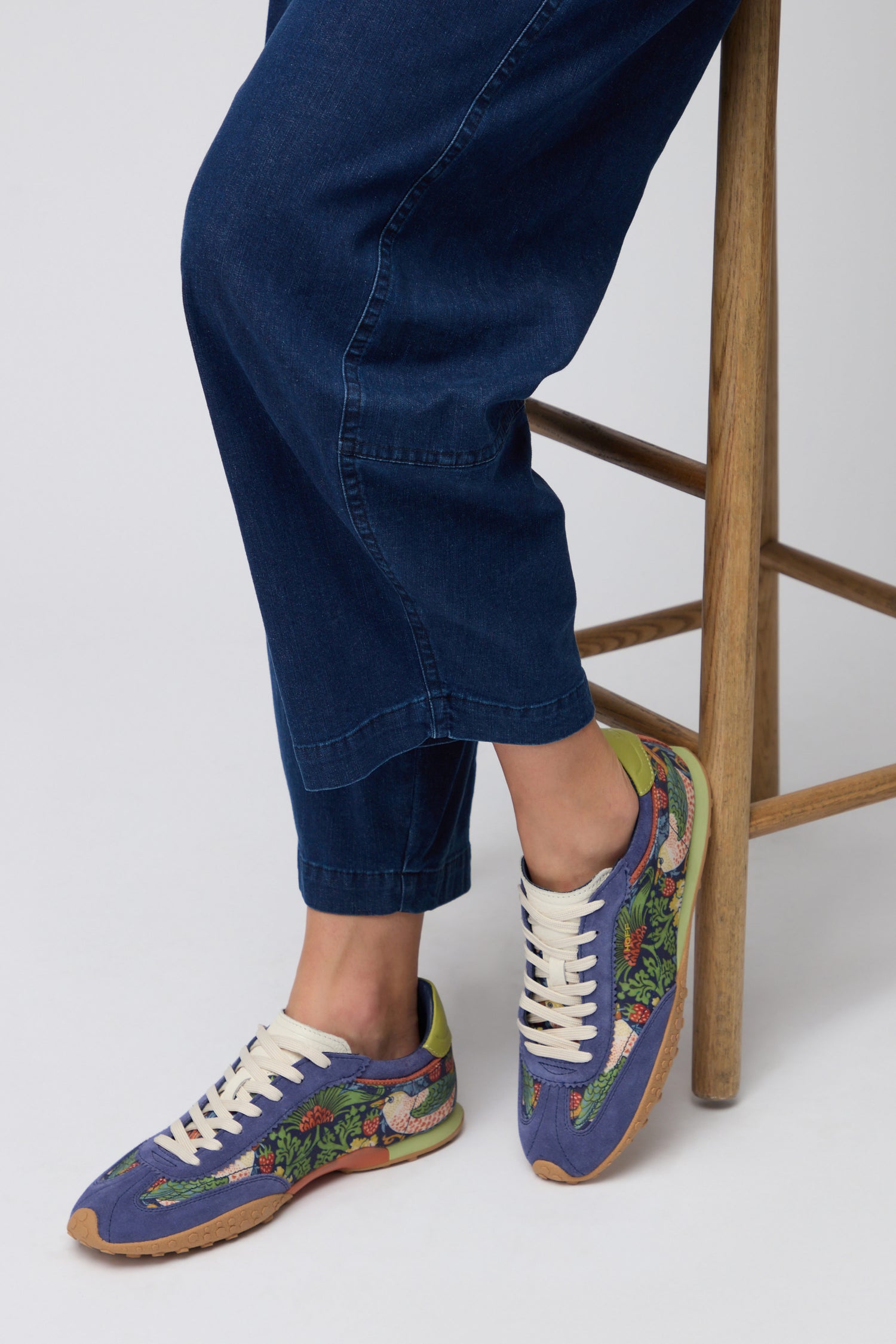 A person wearing blue jeans and Blue William Morris Bridge Trainers sits on a wooden stool against a plain background.