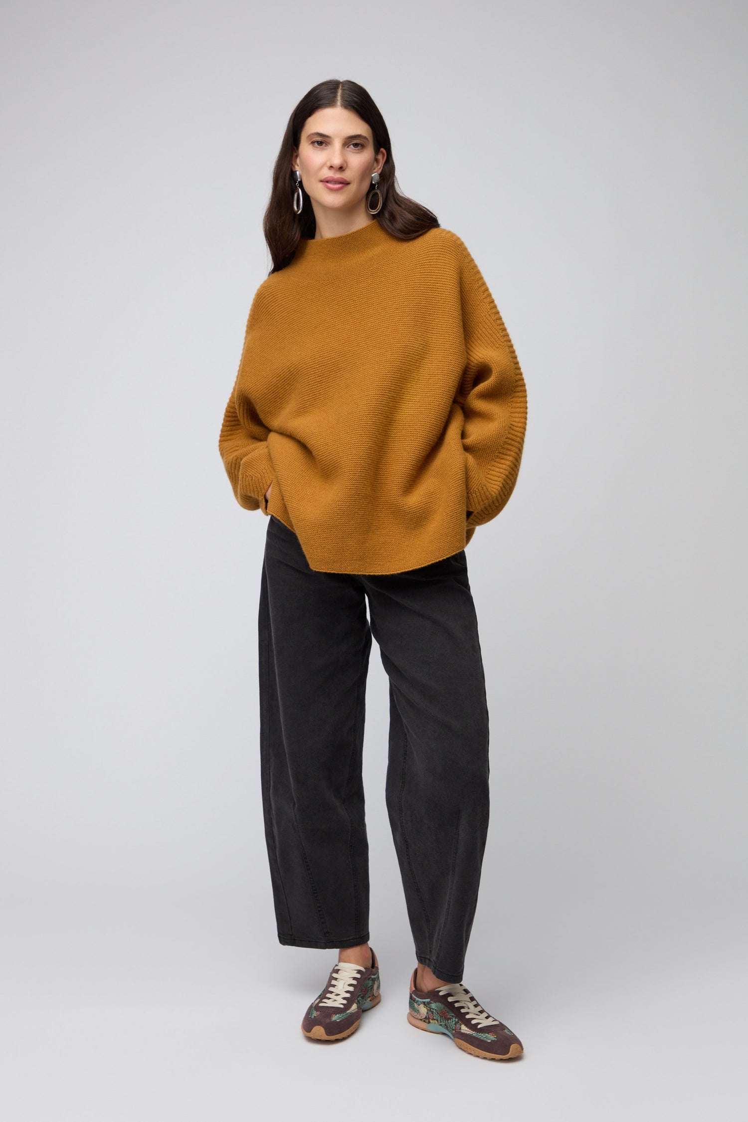 A woman with long dark hair stands against a plain background, wearing a loose mustard sweater, wide-leg black pants, and Brown William Morris Bridge Trainers, looking directly at the camera.