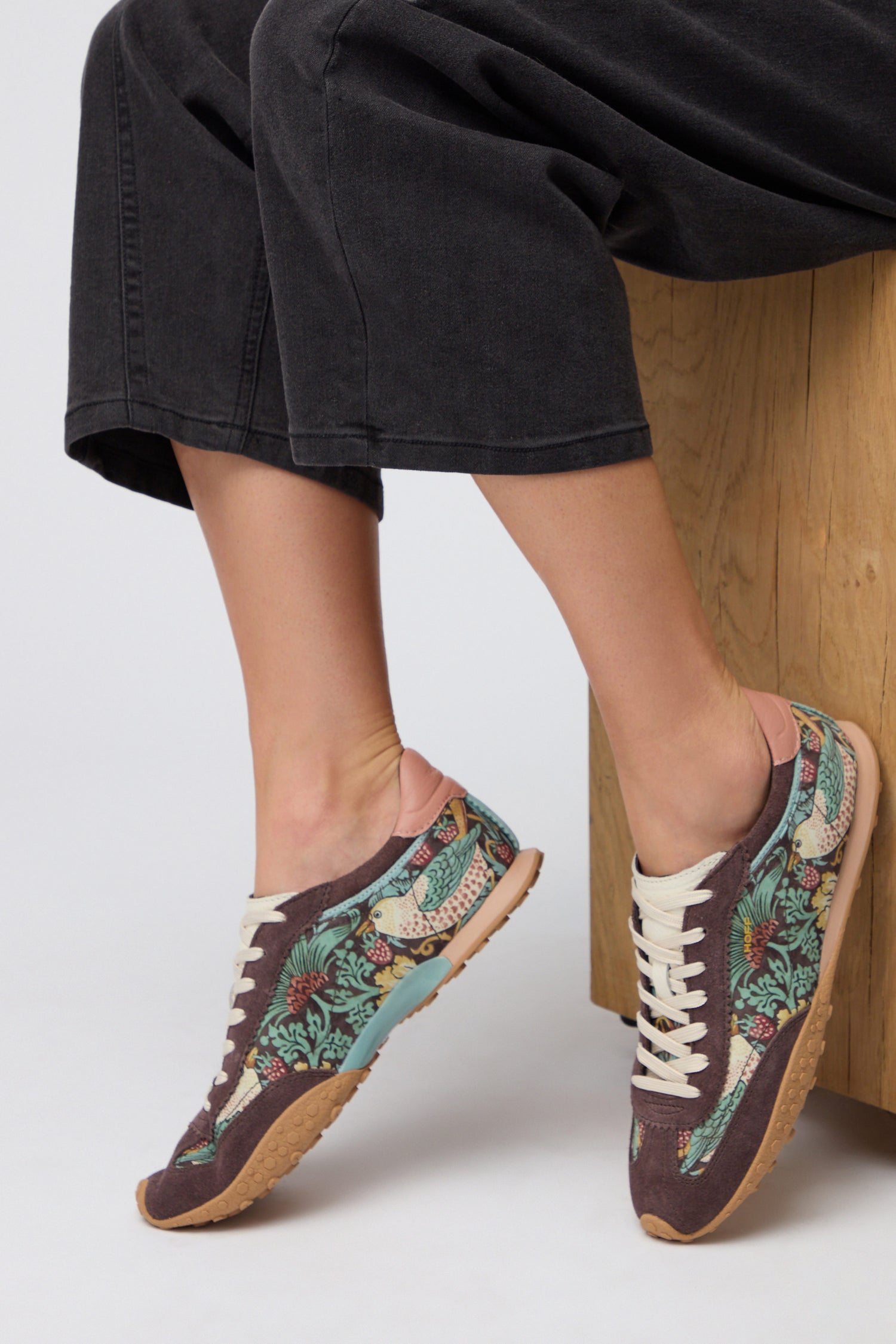 A person wearing Brown William Morris Bridge Trainers with beige laces and black wide-legged pants sits on a wooden bench.