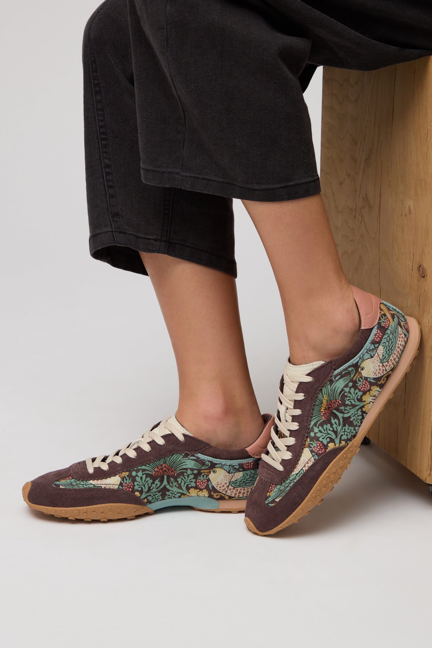 A person wearing Brown William Morris Bridge Trainers—floral-patterned sneakers with white laces and gum soles—sits on a wooden bench.