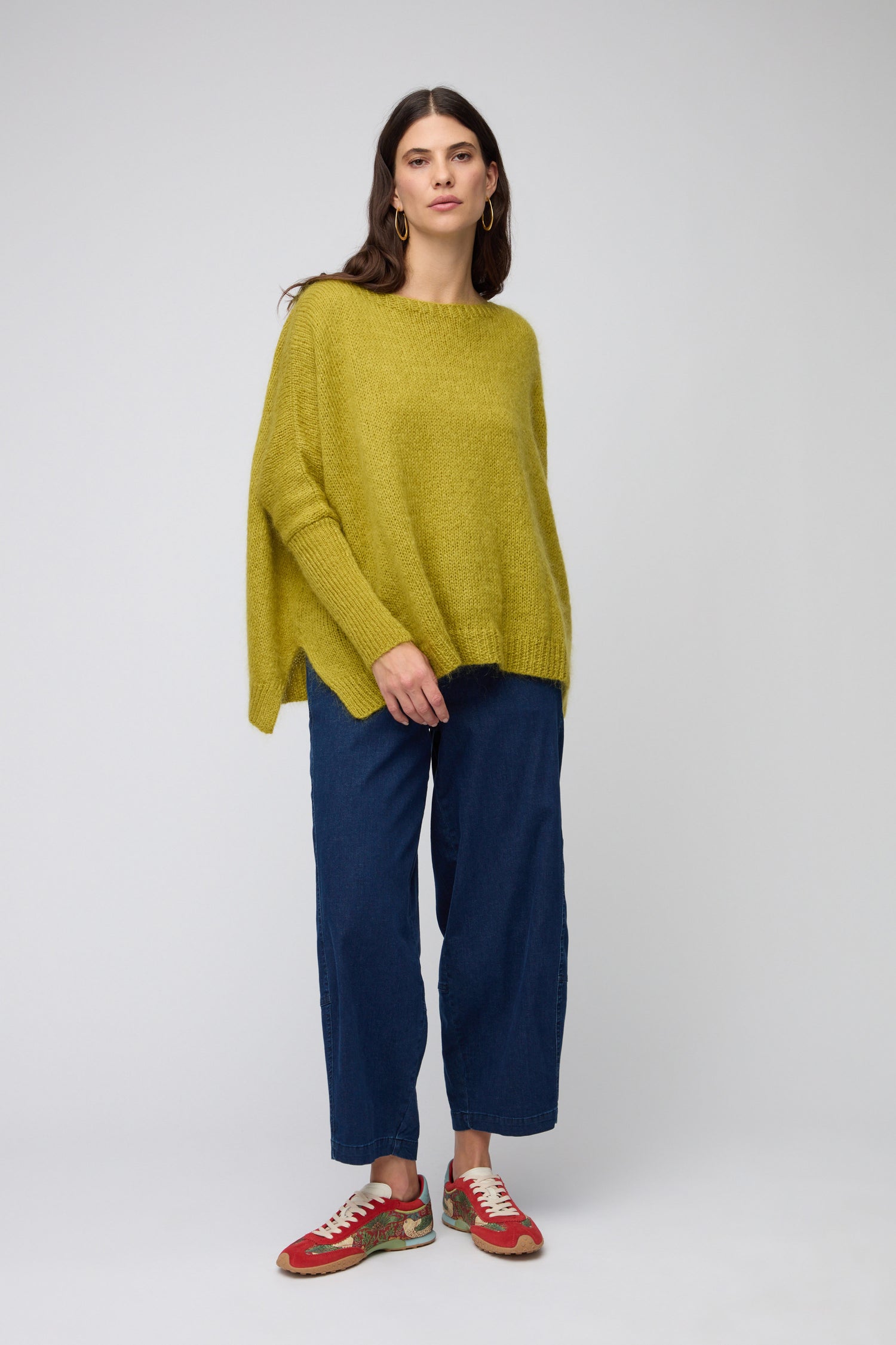 A woman stands against a plain background wearing a loose yellow-green sweater, wide-leg blue jeans, and Red William Morris Bridge Trainers.
