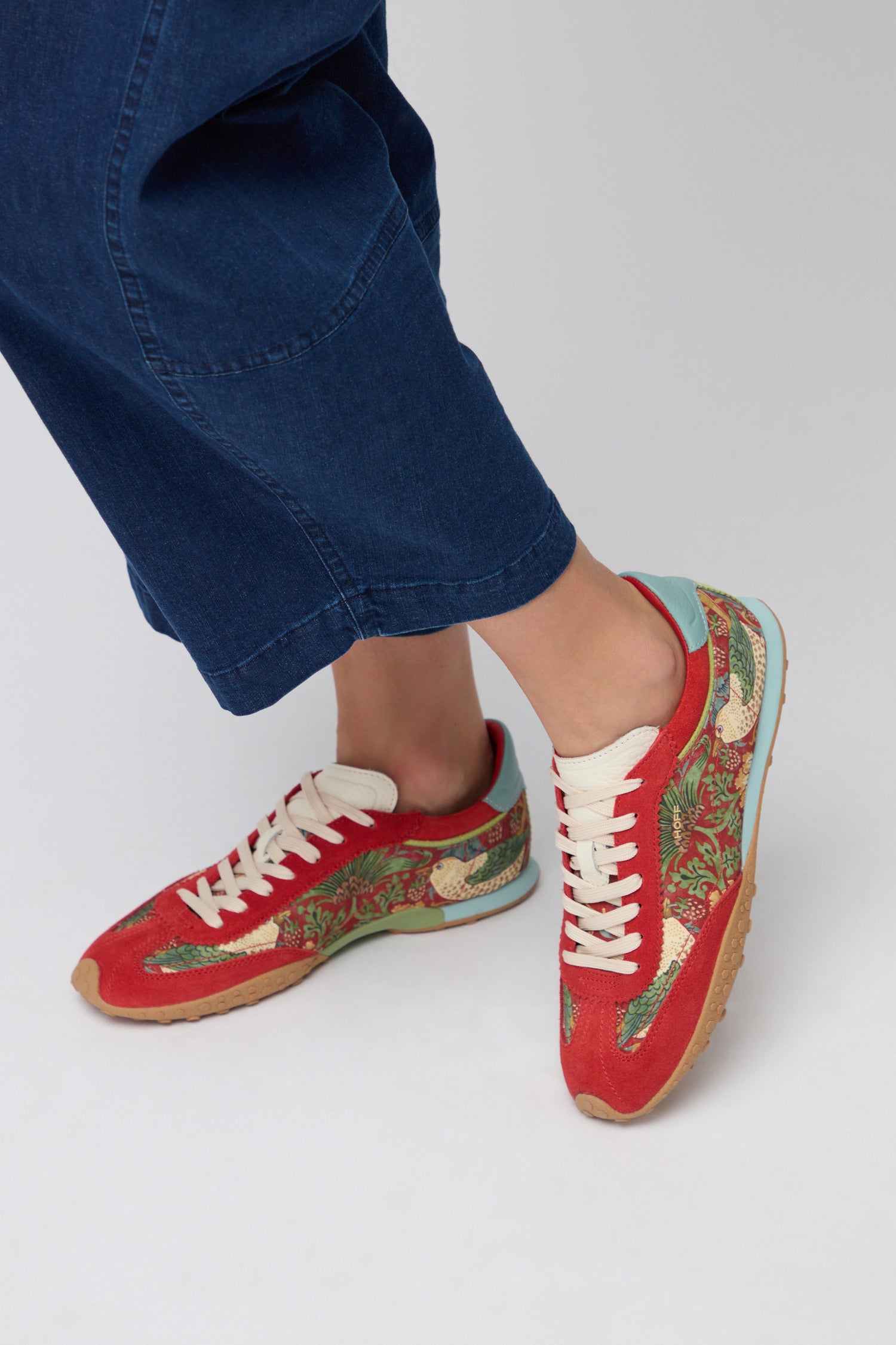 A person wearing blue pants and Red William Morris Bridge Trainers with white laces and a decorative Strawberry Thief print stands on a light gray surface.