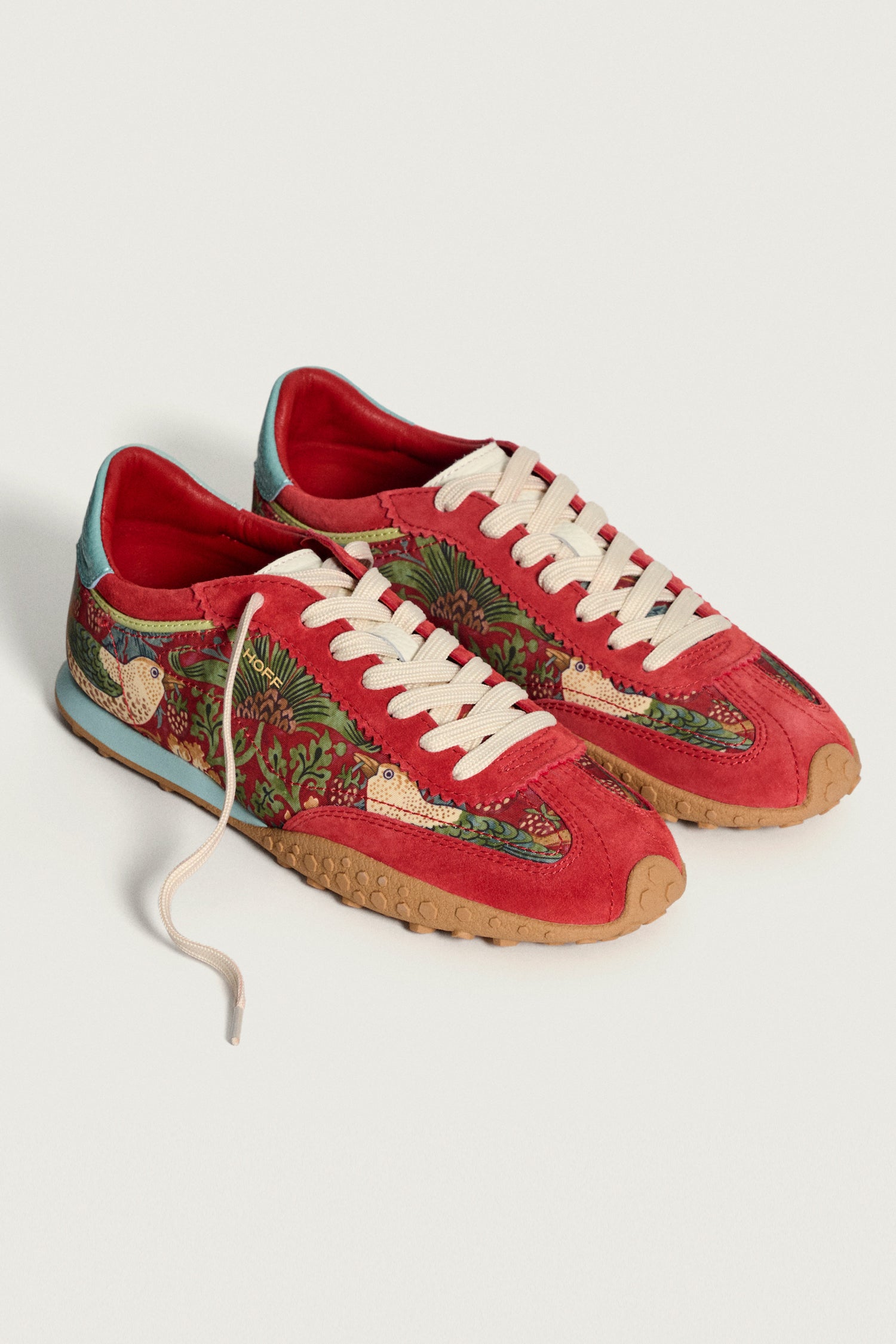 The Red William Morris Bridge Trainers are women’s red low-top leather sneakers with white laces, tan soles, and embroidered floral and bird motifs inspired by the Strawberry Thief print on the sides.