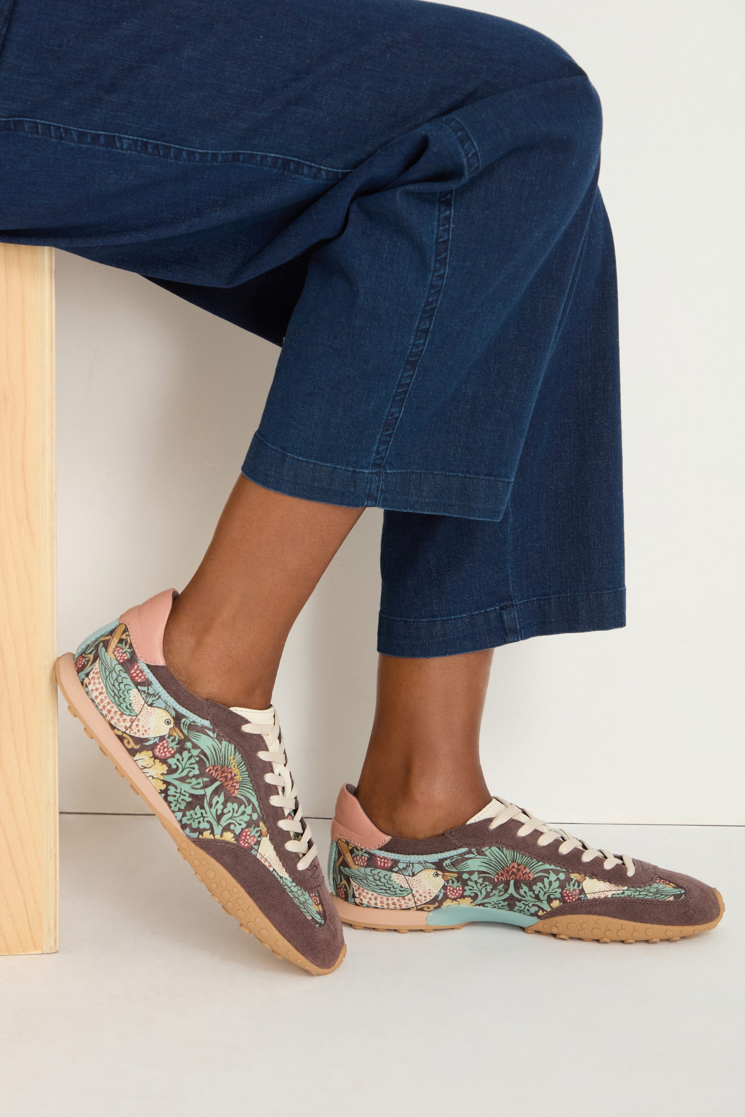 Seated on a light wooden bench against a white background, she wears dark blue cropped pants and William Morris Bridge Trainers with an embroidered tropical pattern.